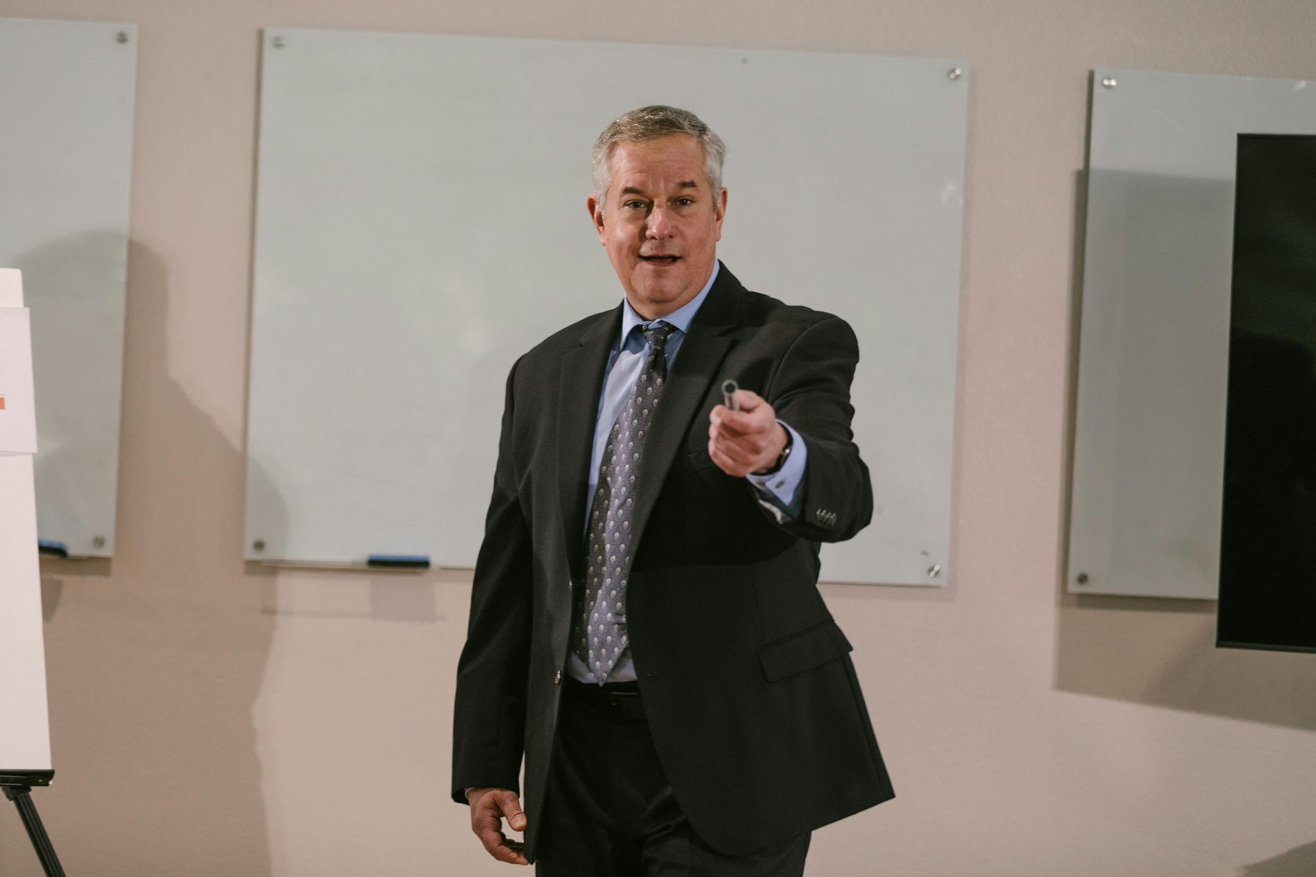 Man in a suit speaking in front of a whiteboard, gesturing in a classroom or meeting room.