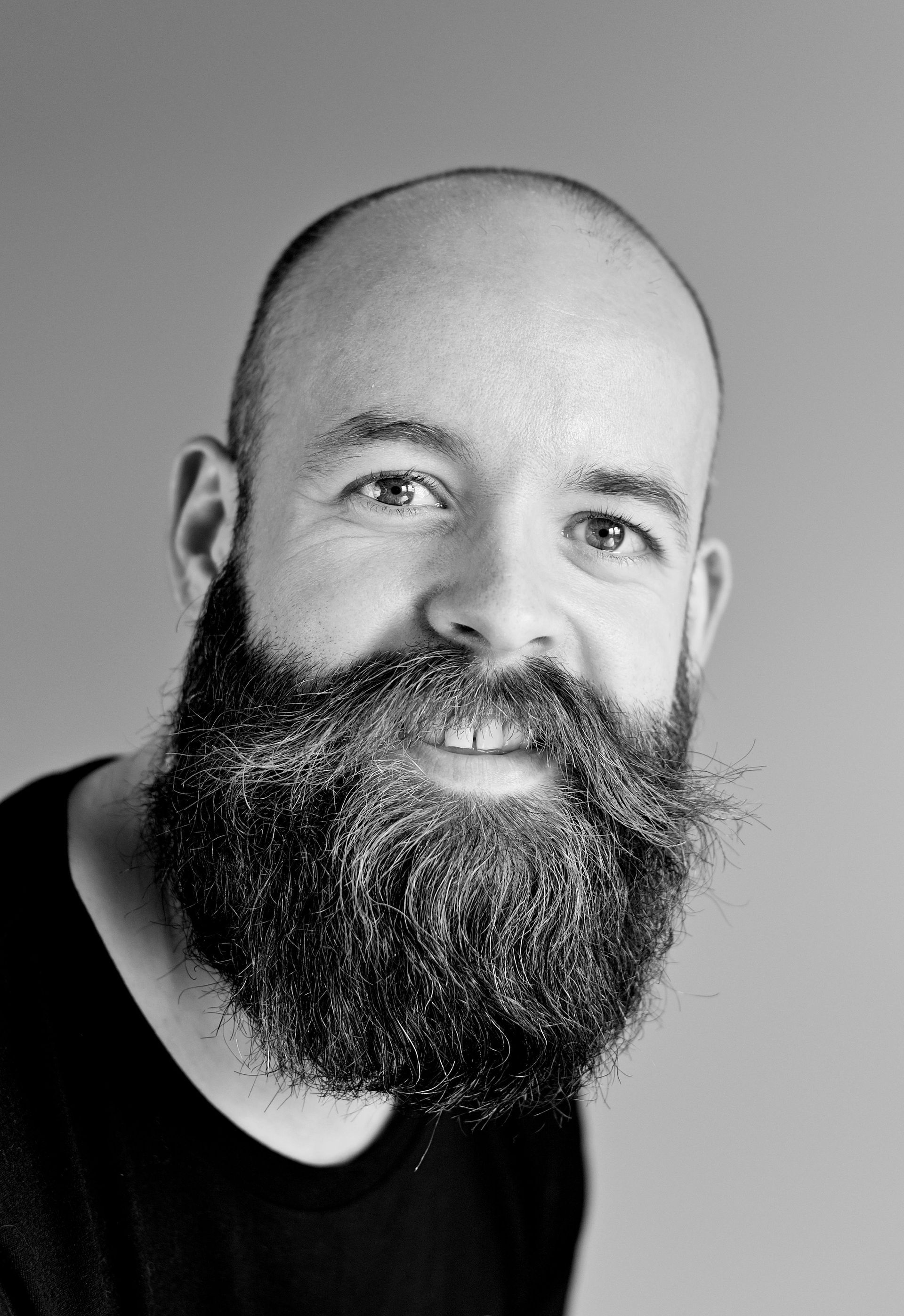 Black-and-white headshot of a smiling bald person with a full beard, wearing a dark shirt.