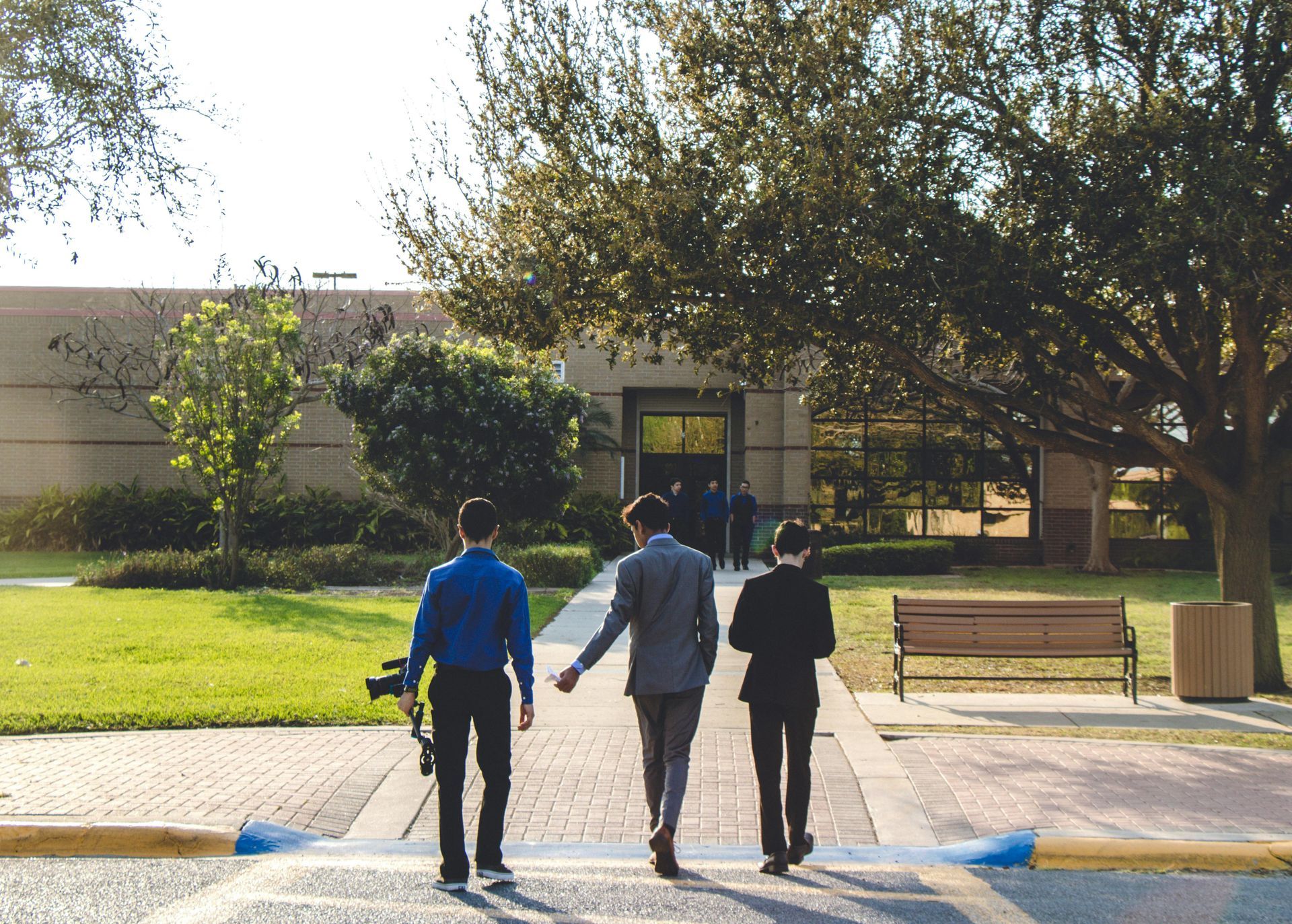 Three people walking on a campus path toward a building, with trees, grass, and a bench nearby.