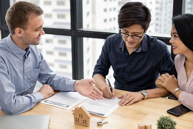 Three people reviewing documents at a table in a bright office, discussing plans with a small model house nearby