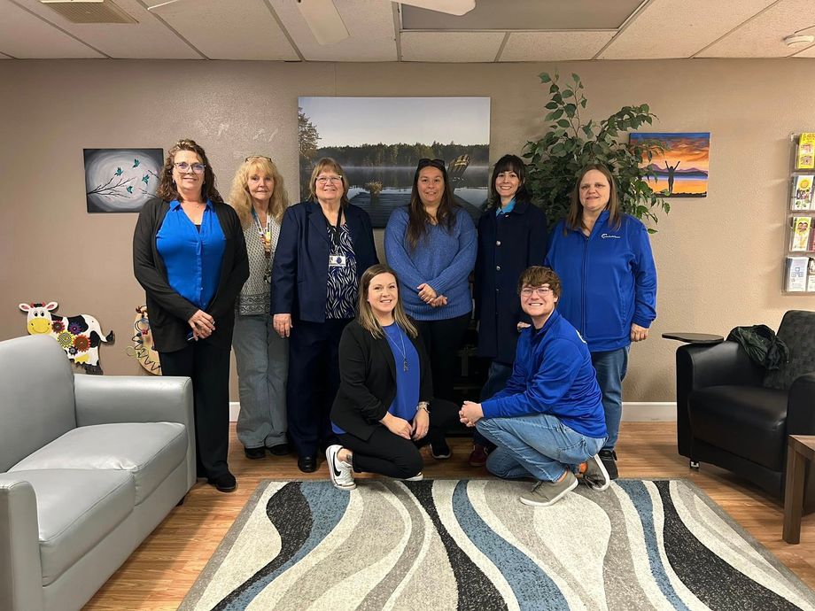 A group of eight people stand and kneel together in an office lobby with blue accents and wall art, smiling for a photo.