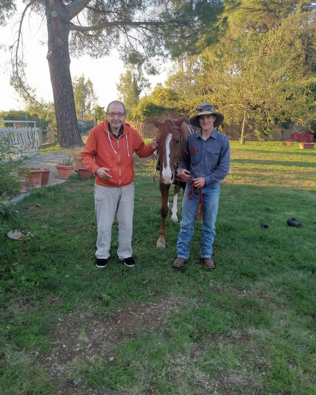 Due persone in piedi con un cavallo in un cortile erboso accanto a un muro di mattoni e ad alberi.