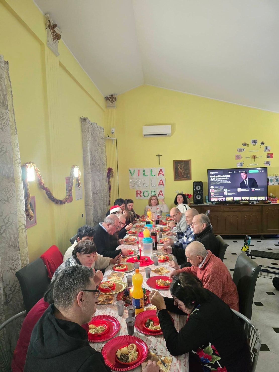 Persone sedute attorno a un lungo tavolo da pranzo in una stanza gialla, che condividono un pasto con piatti rossi e un televisore a parete.