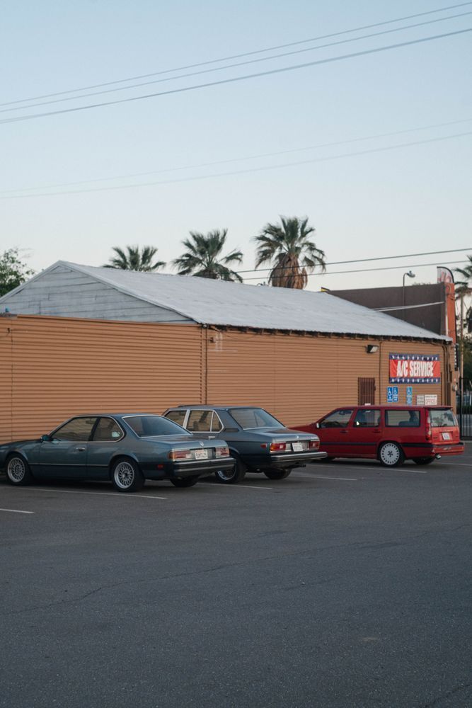 Parked cars in front of a tan building with a white roof and palm trees behind it.