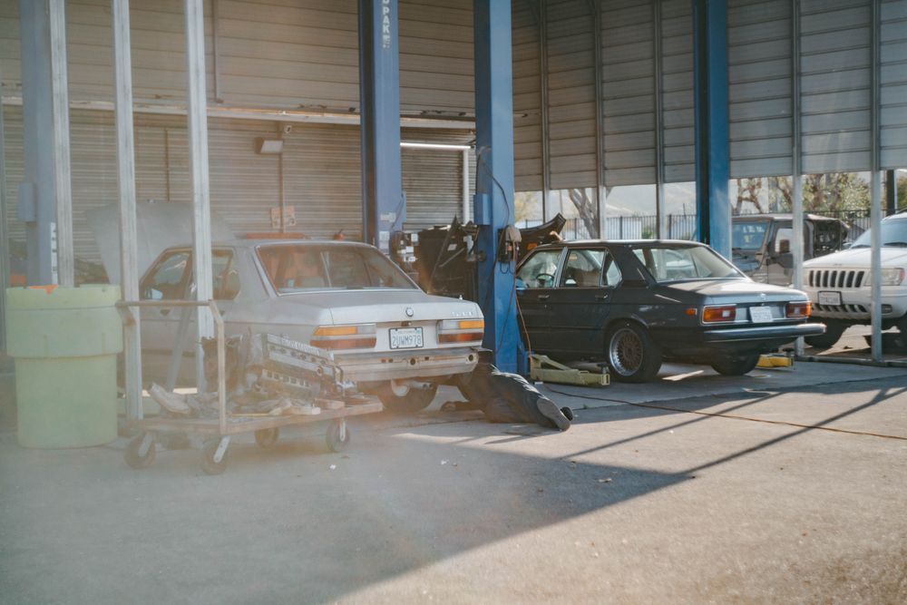 Two parked cars in a garage, with a mechanic working on the white car’s front end.