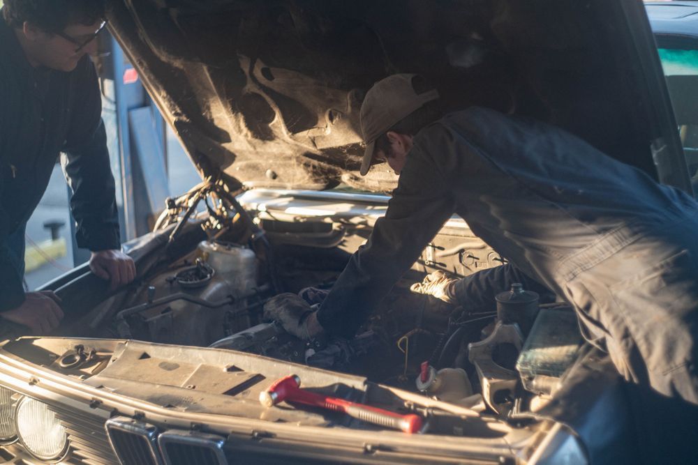 Two people working under a car hood in a garage, with tools on the bumper.