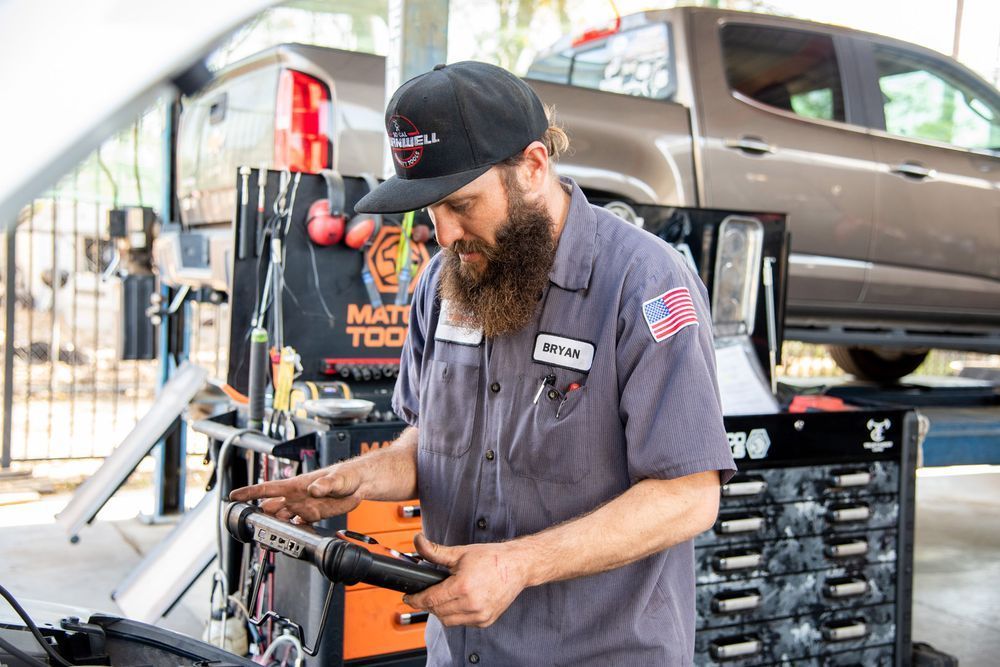 Mechanic working on a car in a garage, holding a power tool beside a lifted vehicle.
