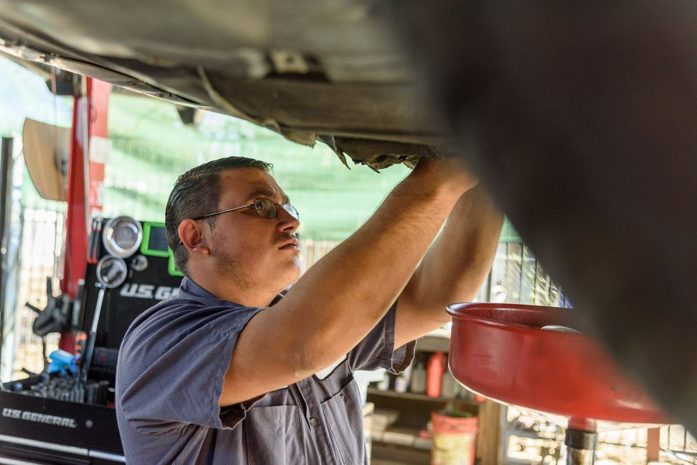 Man tightening equipment under a vehicle, with a red oil pan and workshop tools nearby