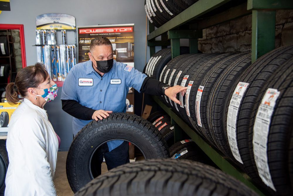 Two masked workers inspect rows of tires in a tire shop.