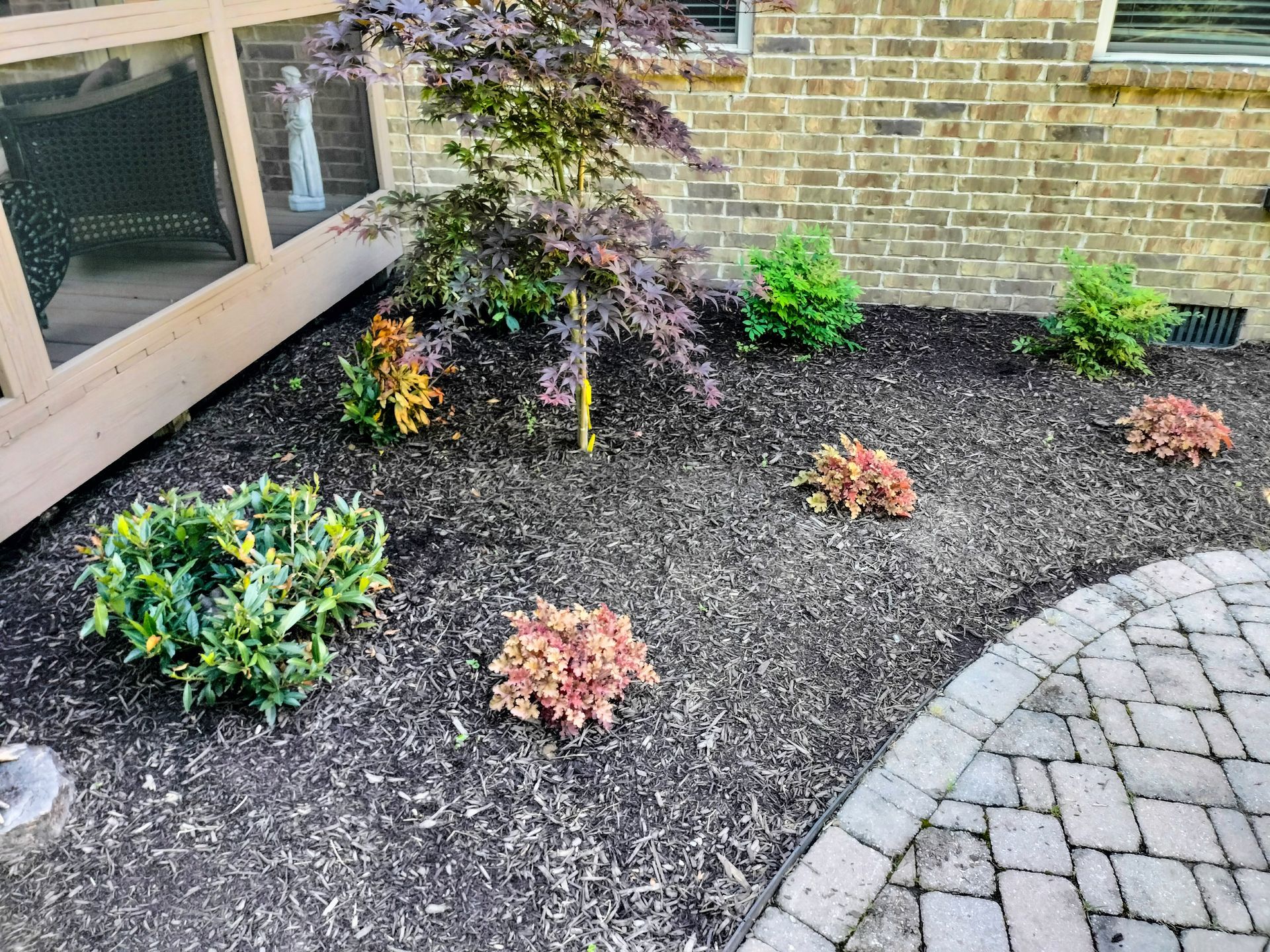 Mulched garden bed with small plants next to a brick wall and a paved patio.
