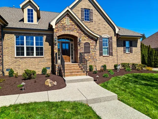 Brick house exterior with front walkway, manicured lawn, and landscaped garden beds