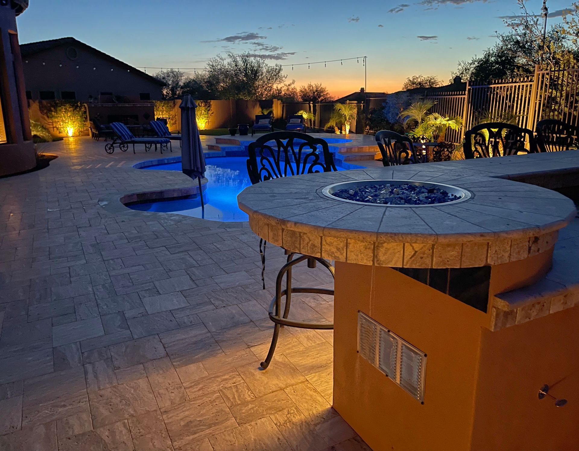 Backyard patio with fire pit, pool, and lighting at dusk.