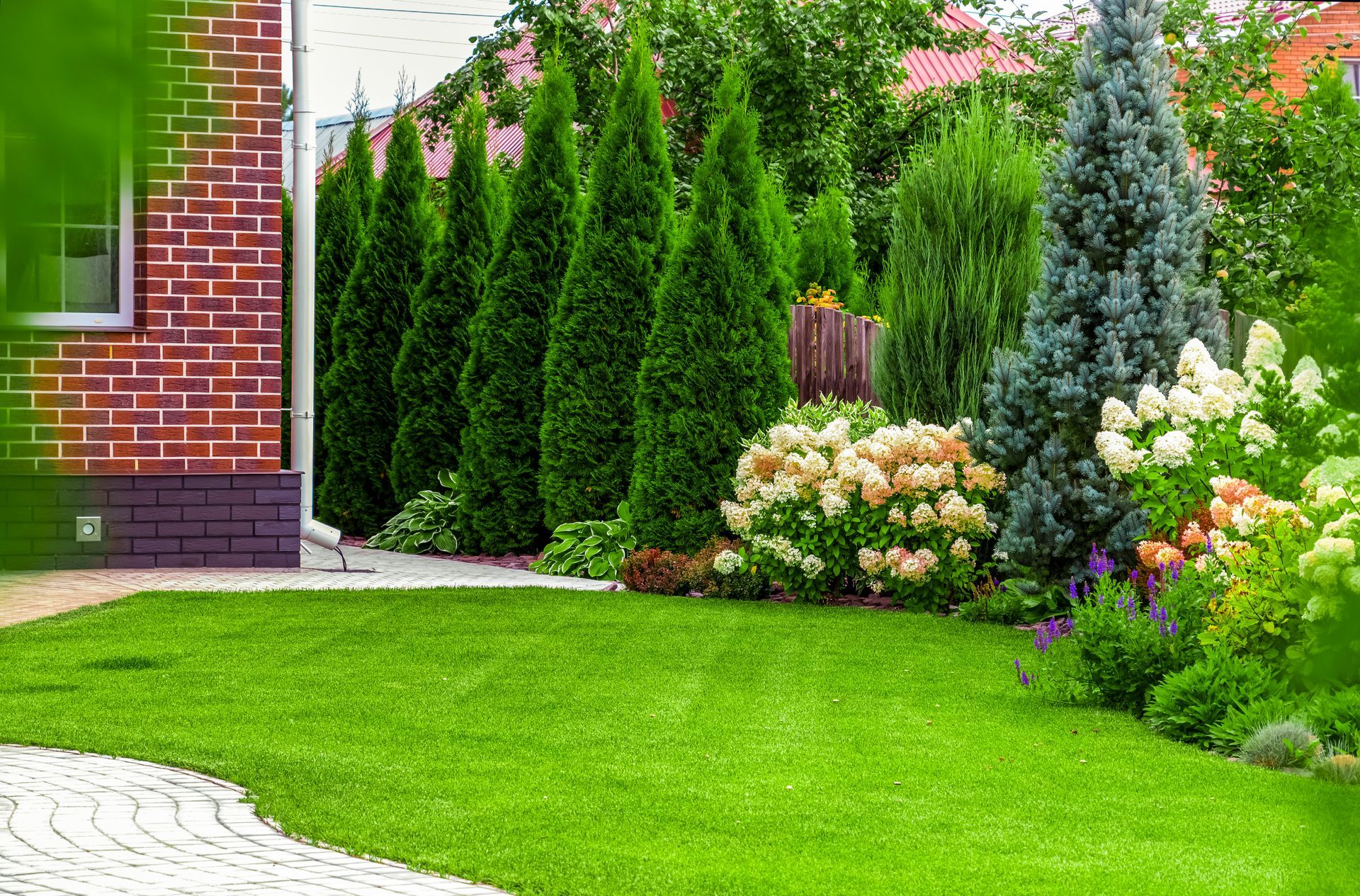Green lawn with evergreen trees and flowering bushes next to a brick building.