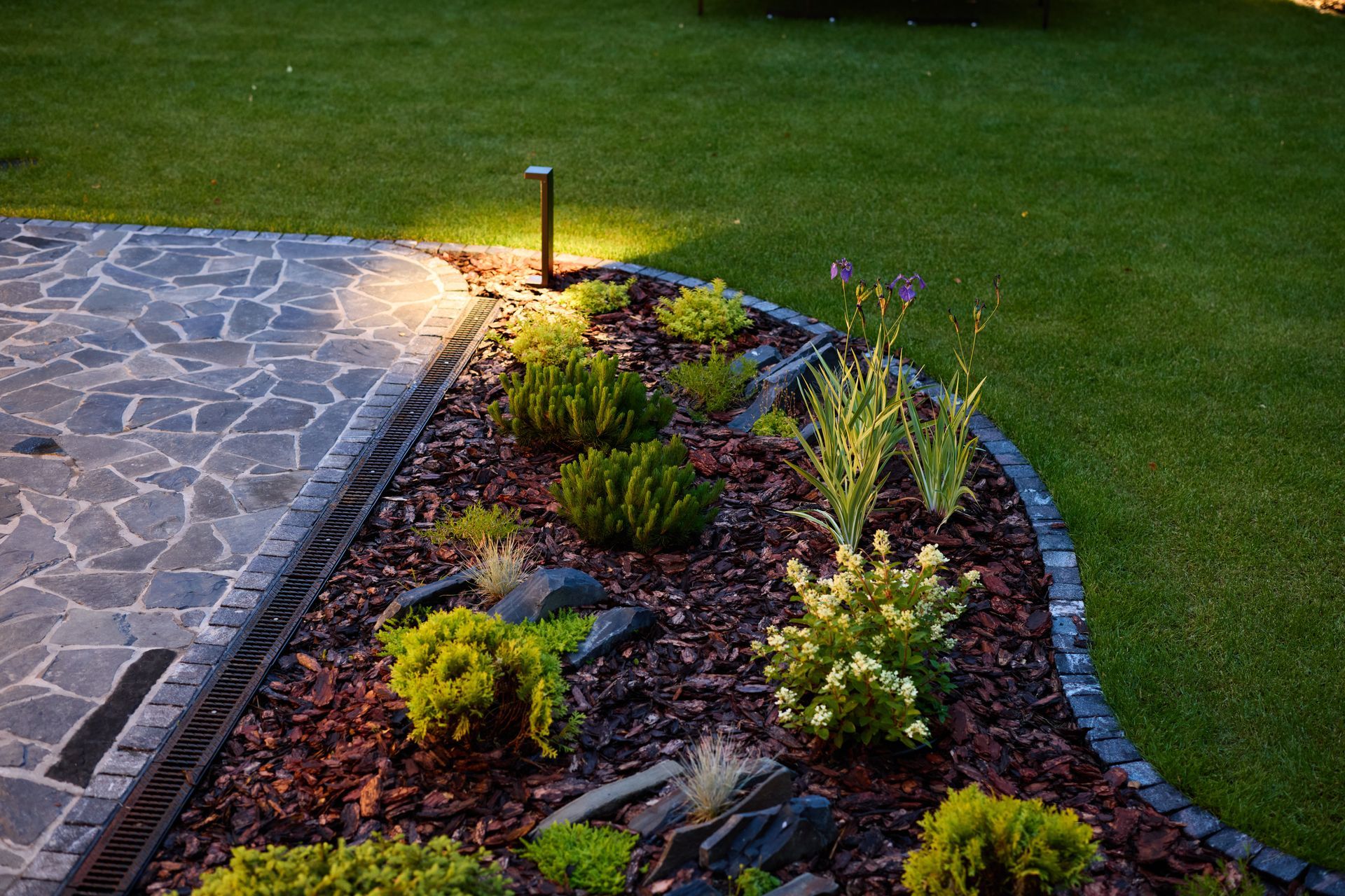 Nighttime landscape with illuminated garden bed beside a stone patio and lawn.