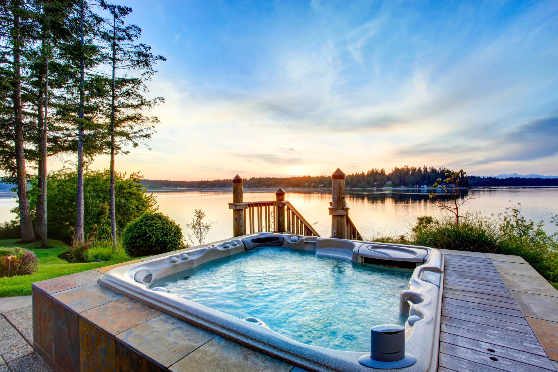 Hot tub overlooking a lake at sunset. Blue water, wooden deck, trees in the background.