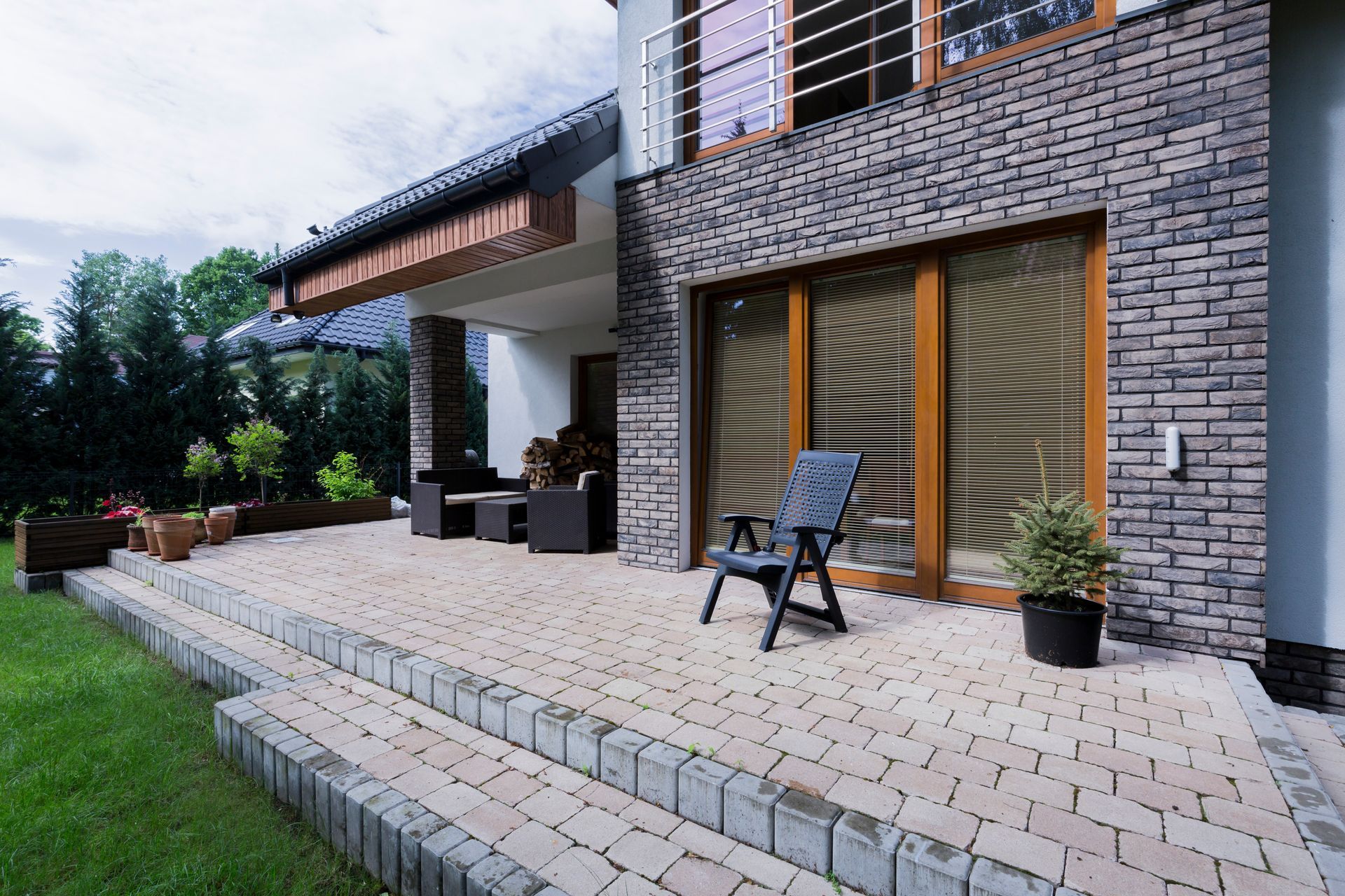 Modern house patio with stone facade, large glass doors, outdoor chairs, and a tiled walkway leading to a lawn