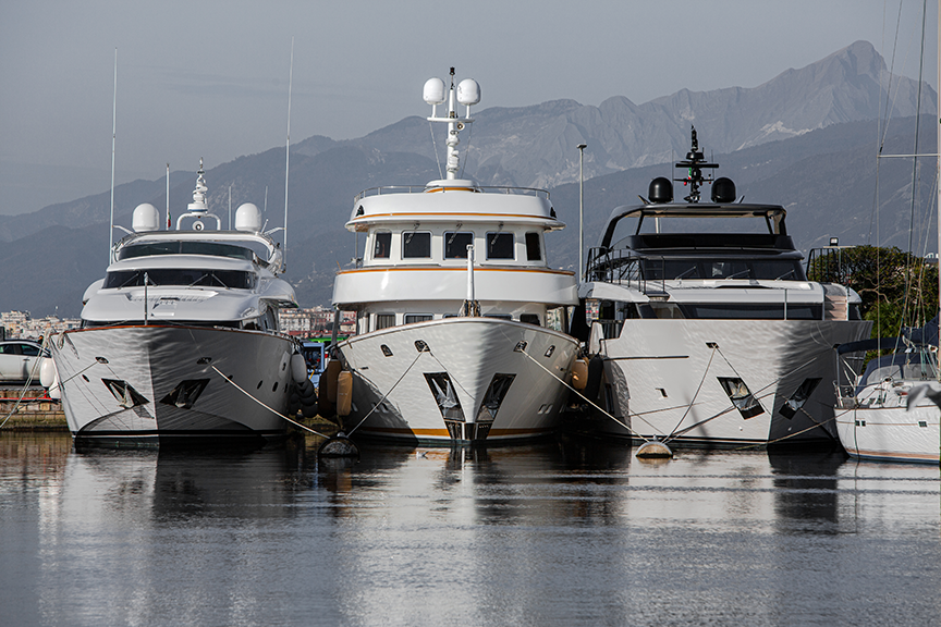 Three luxury yachts docked side by side on calm water with mountains in the background.