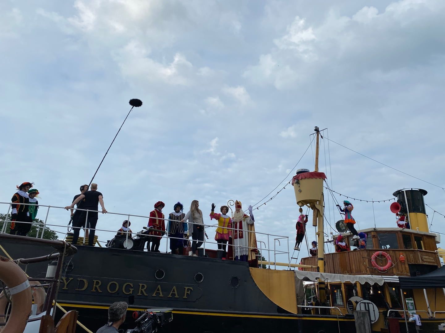 Mensen in feestkleding zwaaien vanaf het dek van een zwarte boot genaamd 'Hydrograaf' onder een bewolkte hemel.
