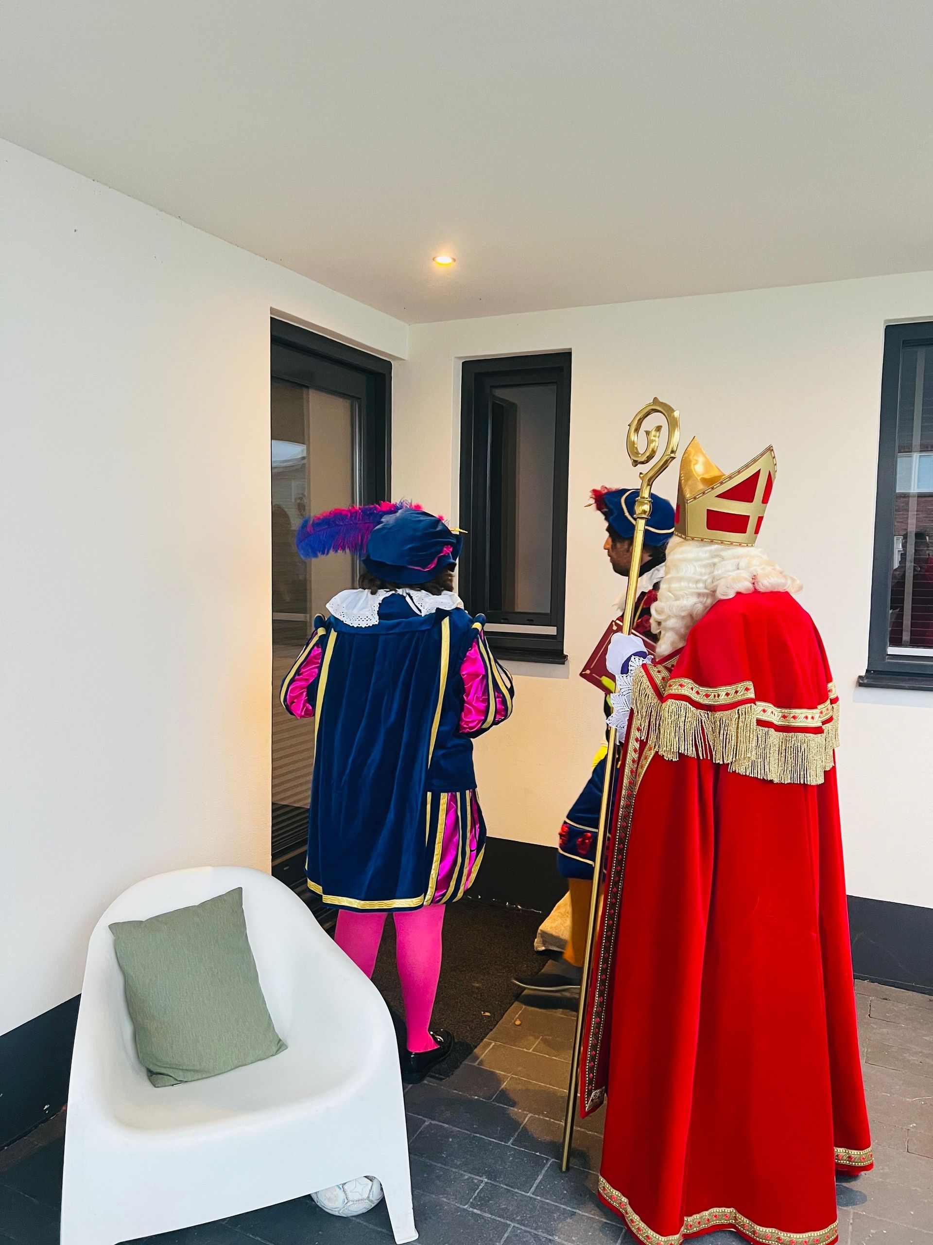 Sinterklaas en twee Pieten in feestelijke, kleurrijke kostuums staan ​​voor een glazen deur op een witte veranda met een witte stoel.