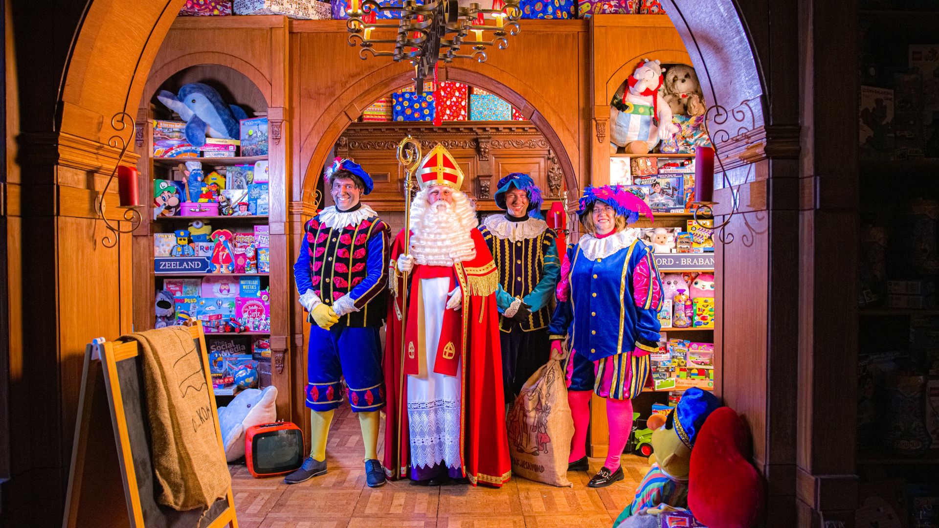 Sinterklaas en zijn drie helpers in kleurrijke kostuums staan ​​in een met speelgoed gevulde kamer die versierd is voor de feestdagen.