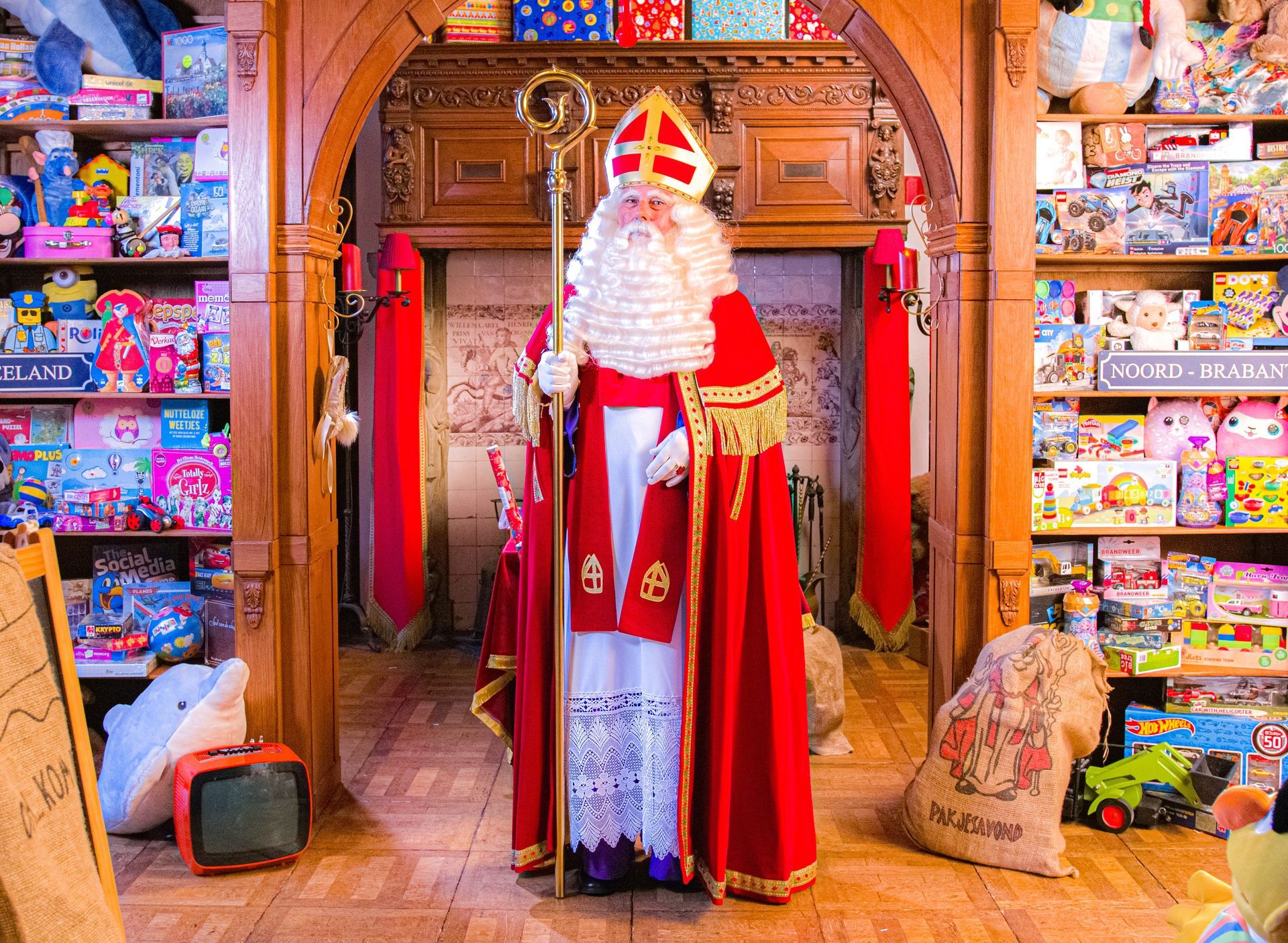 Sint Nicolaas staat in de deuropening van een speelgoedwinkel, met een staf in zijn hand, gekleed in traditionele rood-witte ceremoniële gewaden.