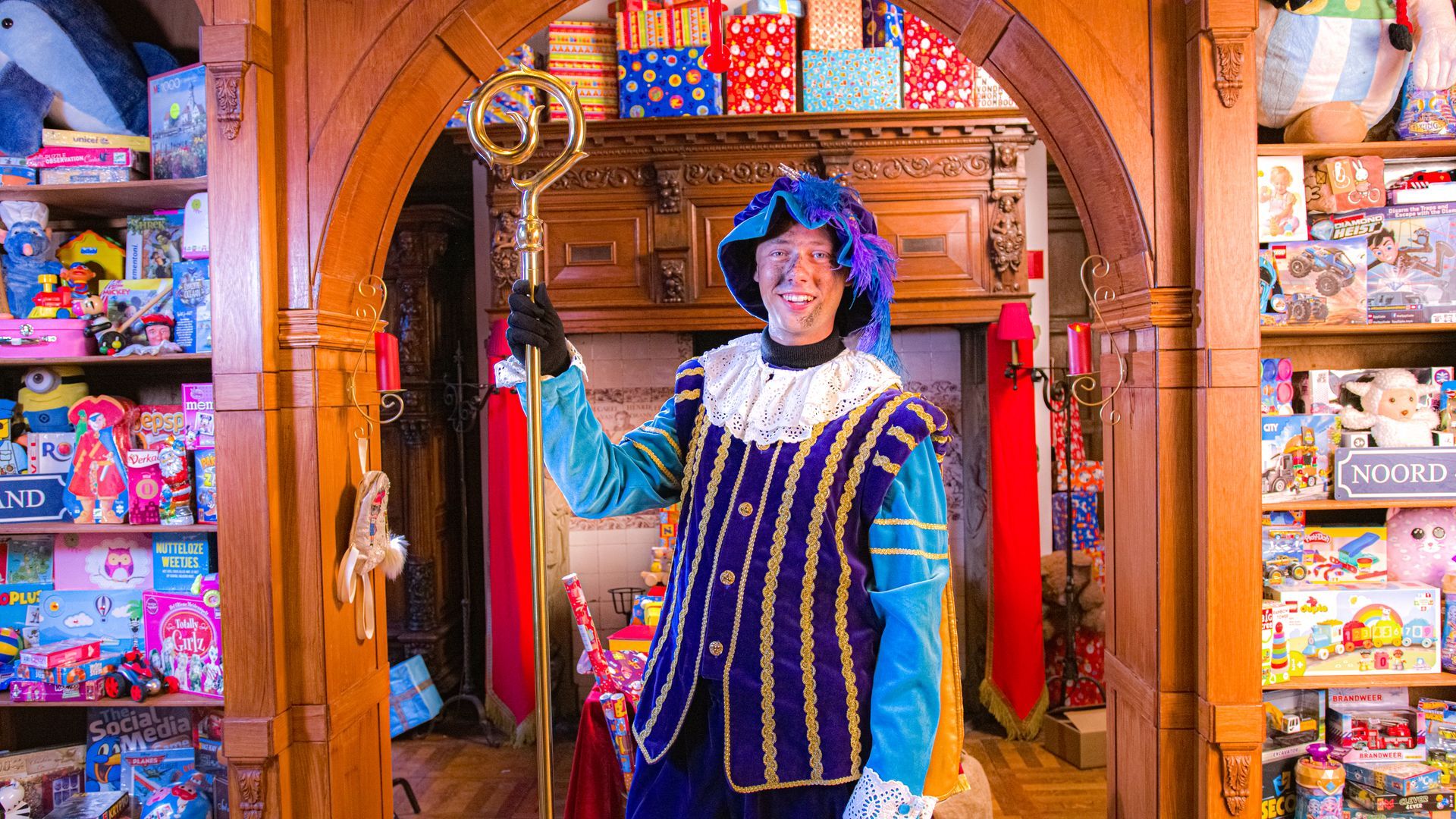 Een persoon, gekleed in een blauw-gouden kostuum in renaissancestijl en met een staf in de hand, staat in een deuropening vol speelgoed.