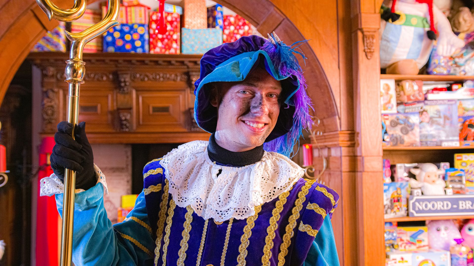 Een persoon gekleed in een feestelijk blauw-goud kostuum met een gevederde hoed en een staf, staat in een kamer vol speelgoed.