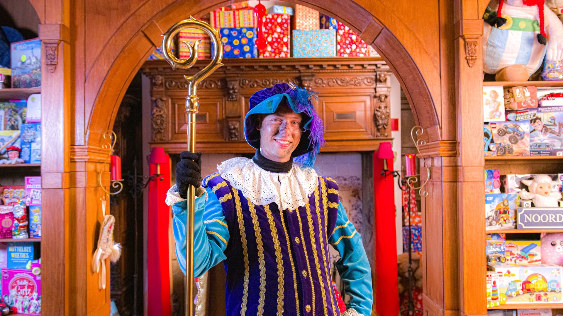 Een persoon in een kleurrijk kostuum en hoed in renaissancestijl houdt een gouden staf vast in een kamer vol geschenkdozen.