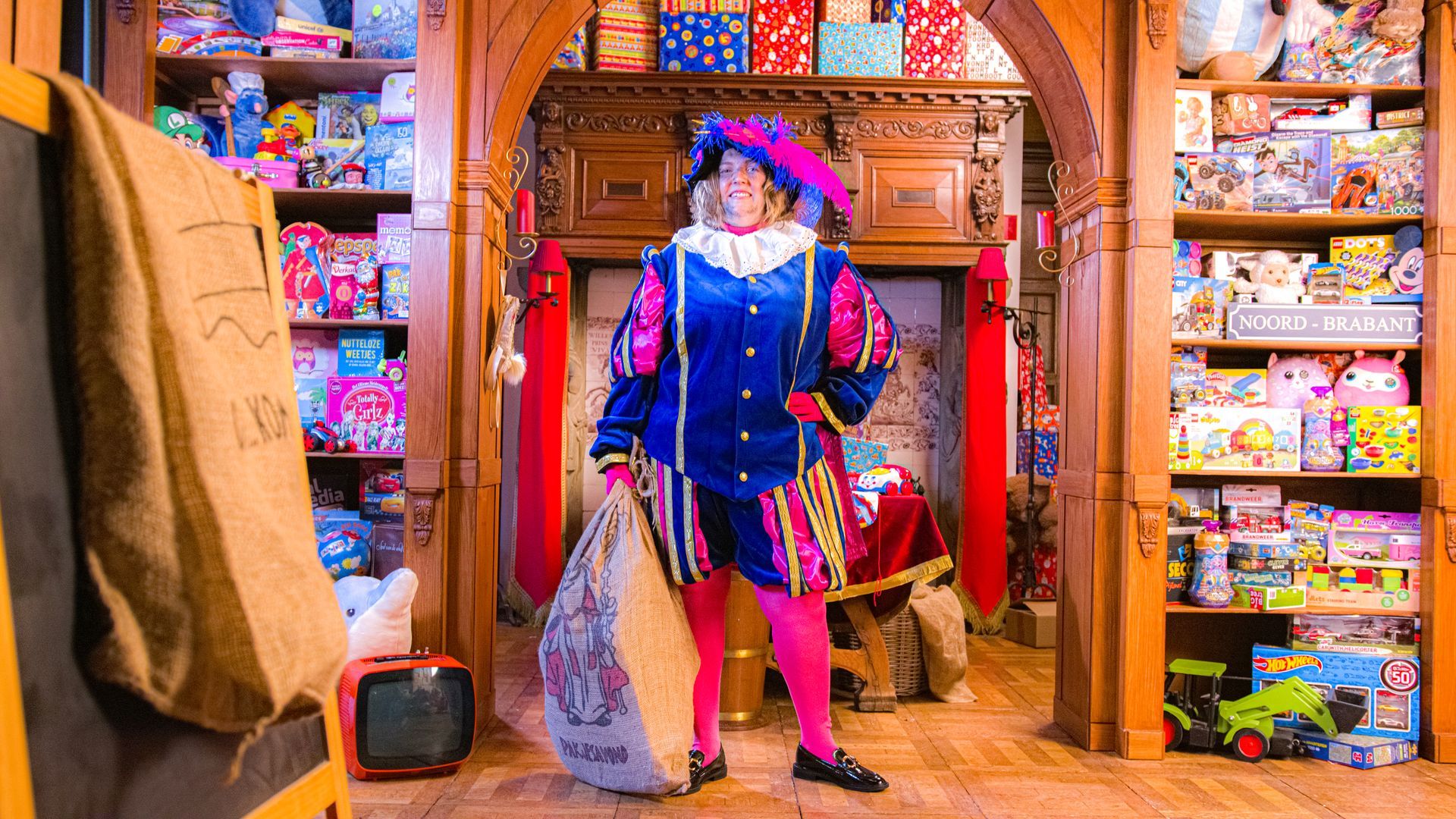 Een persoon in een kleurrijk koninklijk kostuum en hoed staat in een kamer vol speelgoed en houdt een grote stoffen zak vast.