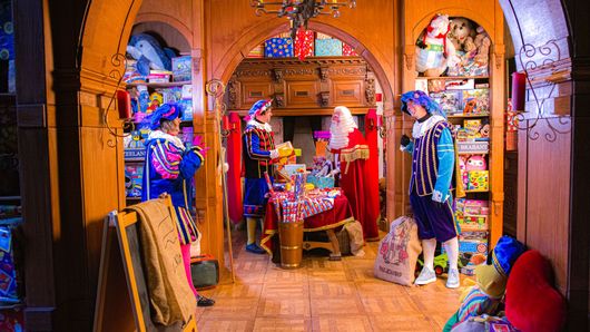 De Kerstman en drie helpers in kleurrijke traditionele kleding staan ​​in een versierde, met speelgoed gevulde houten kamer.