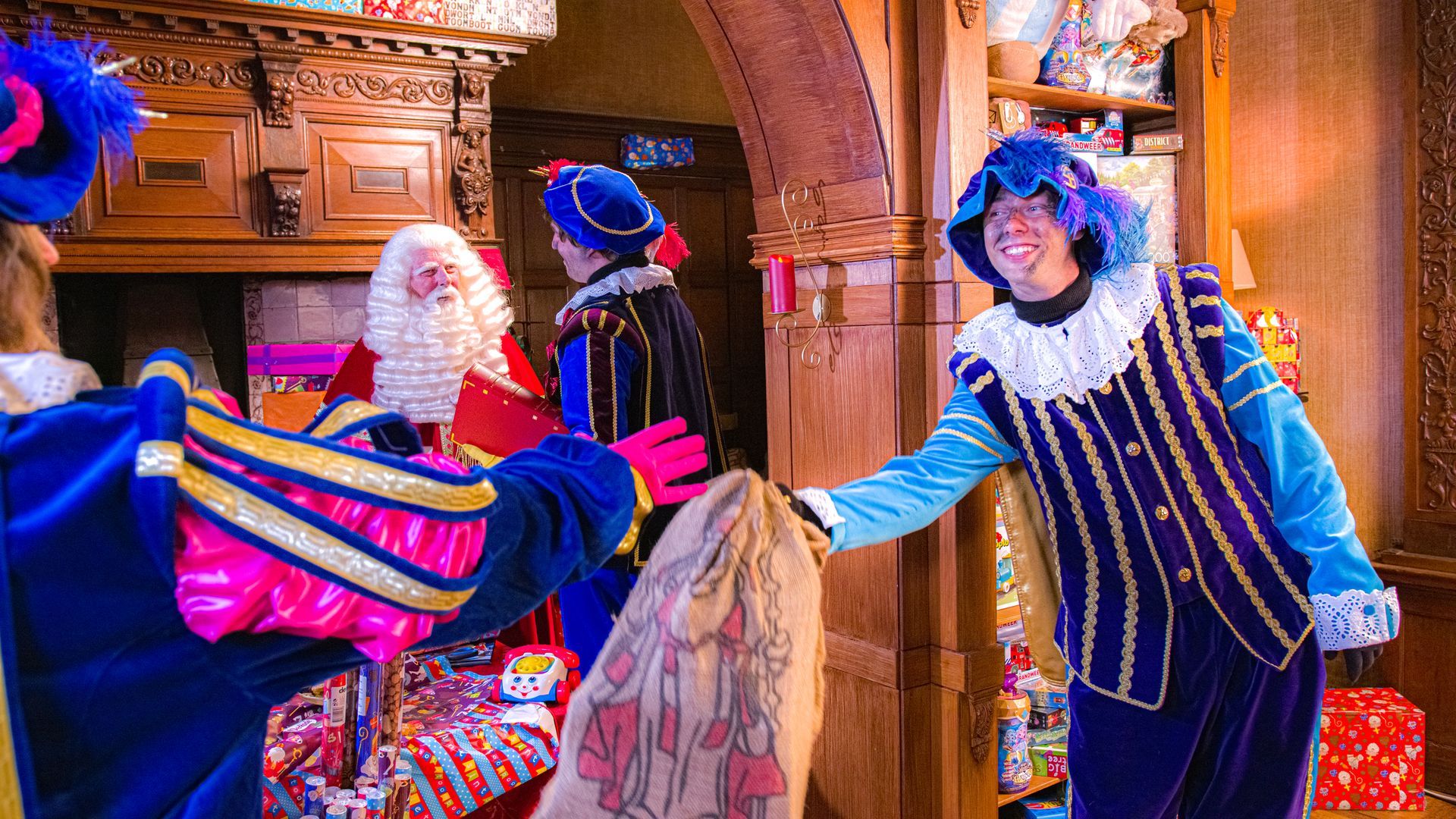 Sinterklaas en zijn helpers in traditionele blauw-gouden kostuums vermaken zich in een kamer met cadeaus en een open haard.