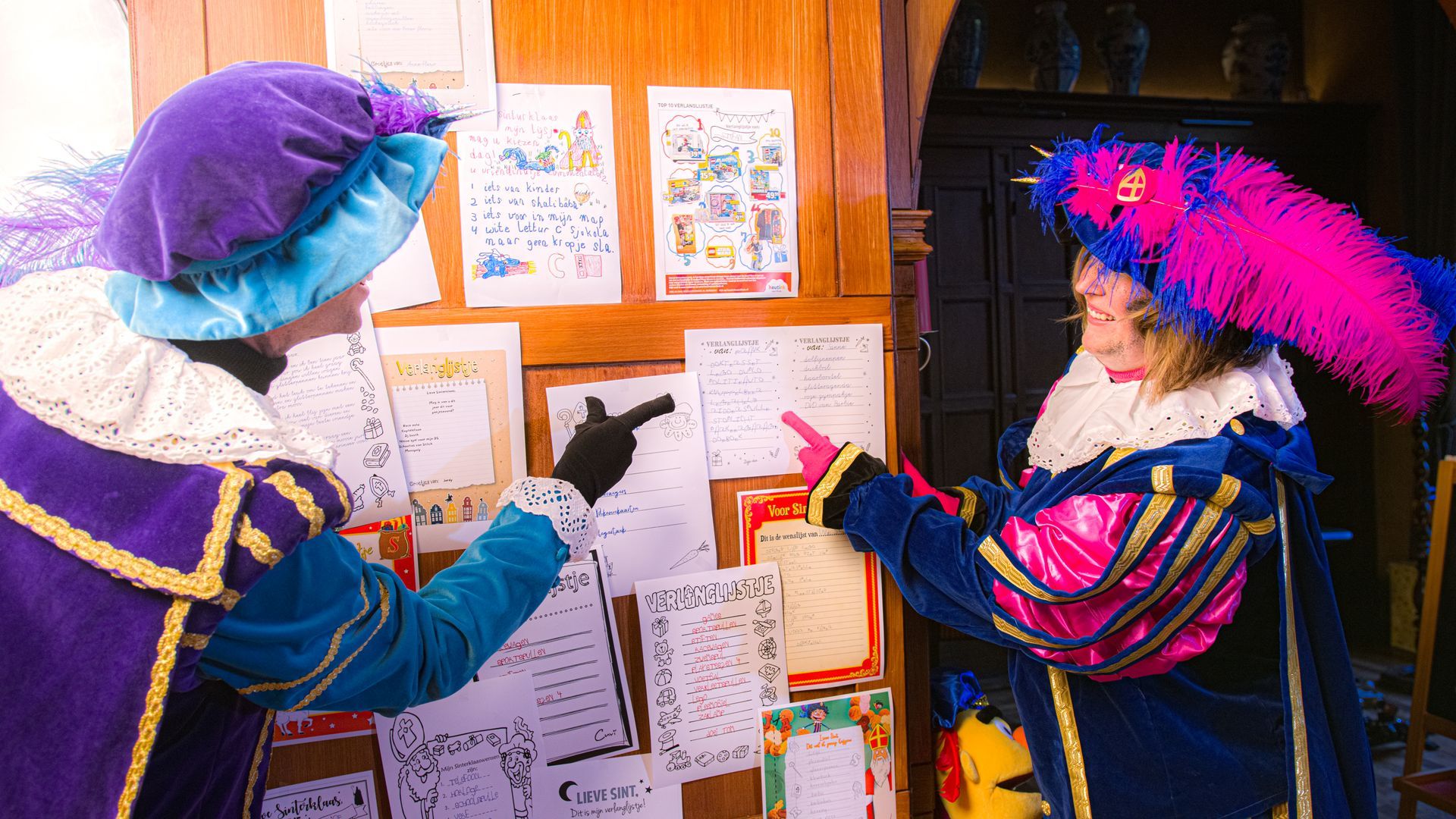 Twee figuren in uitbundige paarse en blauwe historische kostuums en gevederde hoeden wijzen naar papieren die op een houten plank zijn vastgespeld.