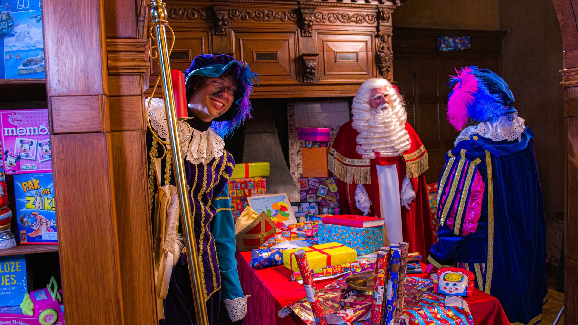 Sinterklaas en twee verklede helpers, omringd door cadeaus in een feestelijke, rustieke binnenomgeving.