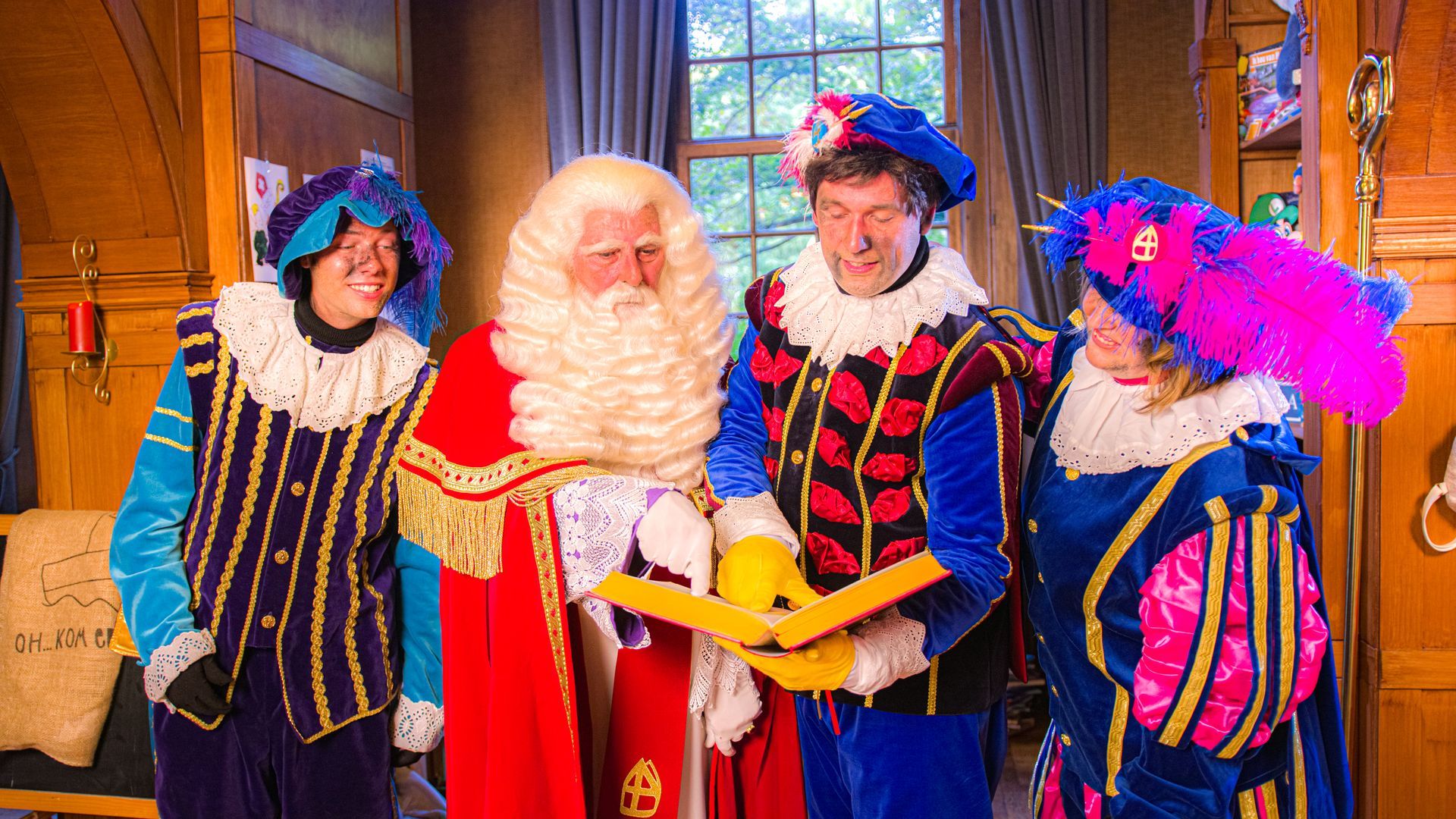 Sinterklaas en zijn drie helpers, gekleed in traditionele, felgekleurde kostuums in renaissancestijl, bekijken binnen een open boek.