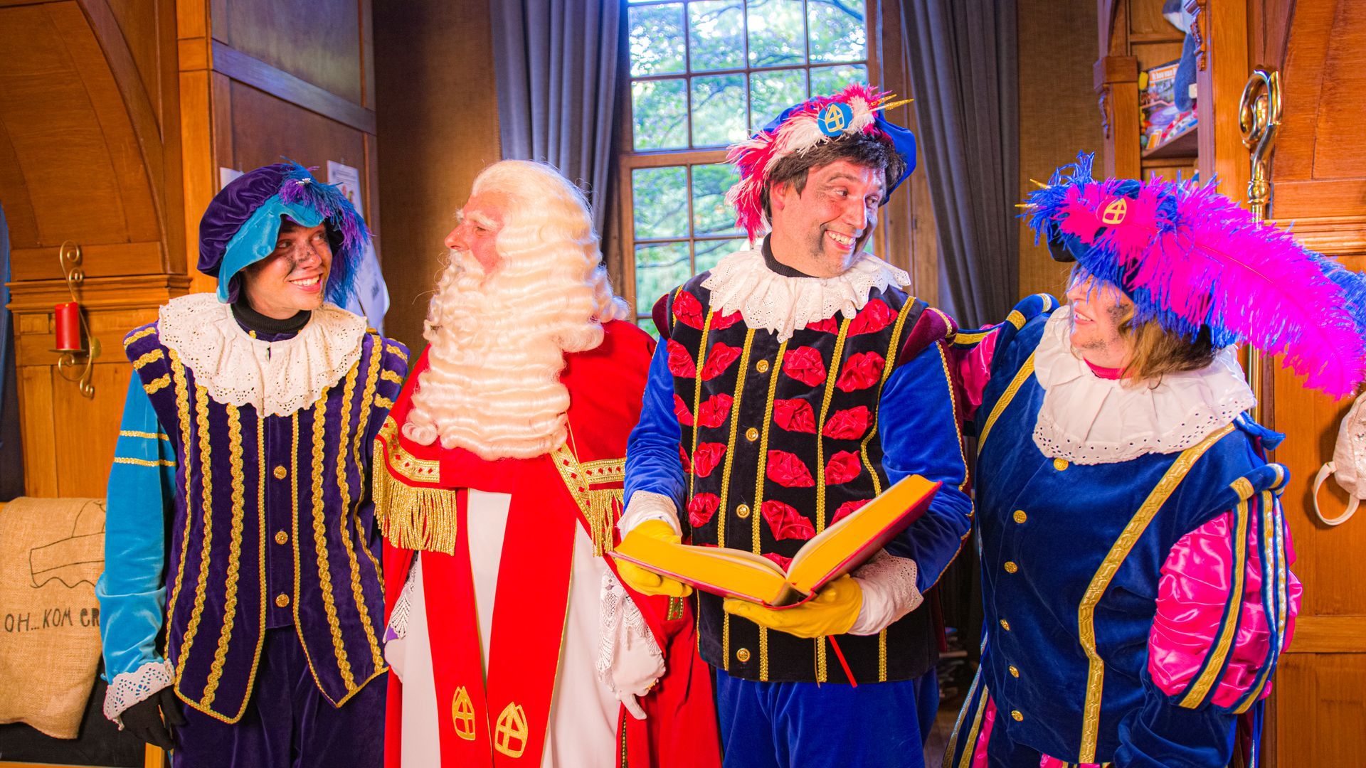 Sinterklaas en drie figuren in uitbundige 17e-eeuwse kostuums staan ​​binnen, glimlachend en in gesprek.