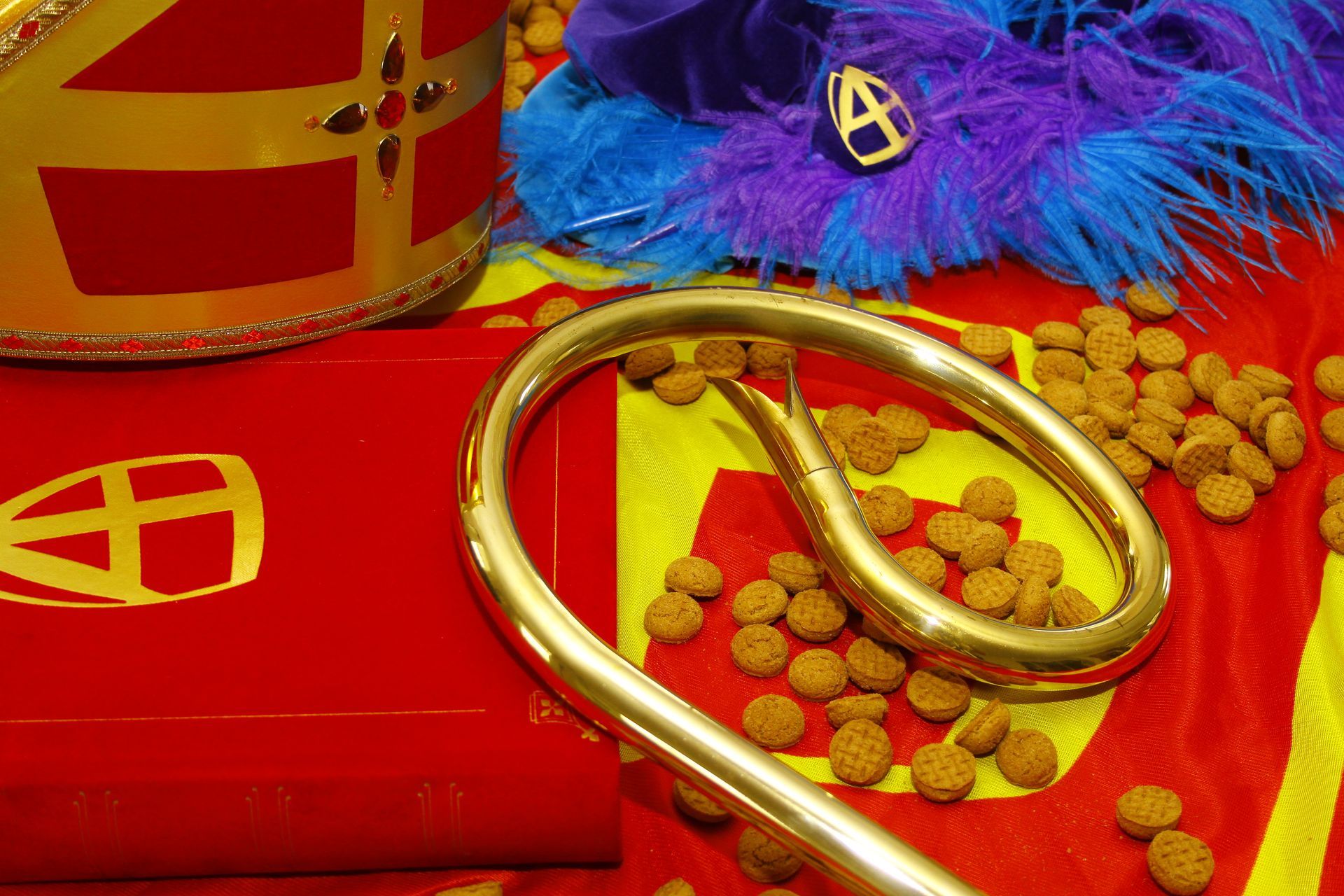 Accessoires voor Sinterklaas: een mijter, een gouden staf, een rood boek en verspreide peperkoekjes (pepernoten) op een stuk stof.