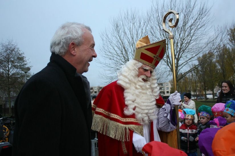 Een man in een zwarte jas glimlacht naar Sint Nicolaas, die een rood gewaad, een witte baard en een mijter draagt ​​en een gouden staf vasthoudt.