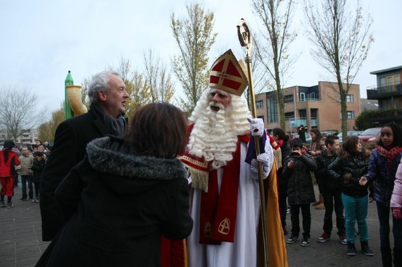Een figuur verkleed als Sinterklaas met staf en mijter heeft een interactie met een persoon in een zwarte jas in de buitenlucht.