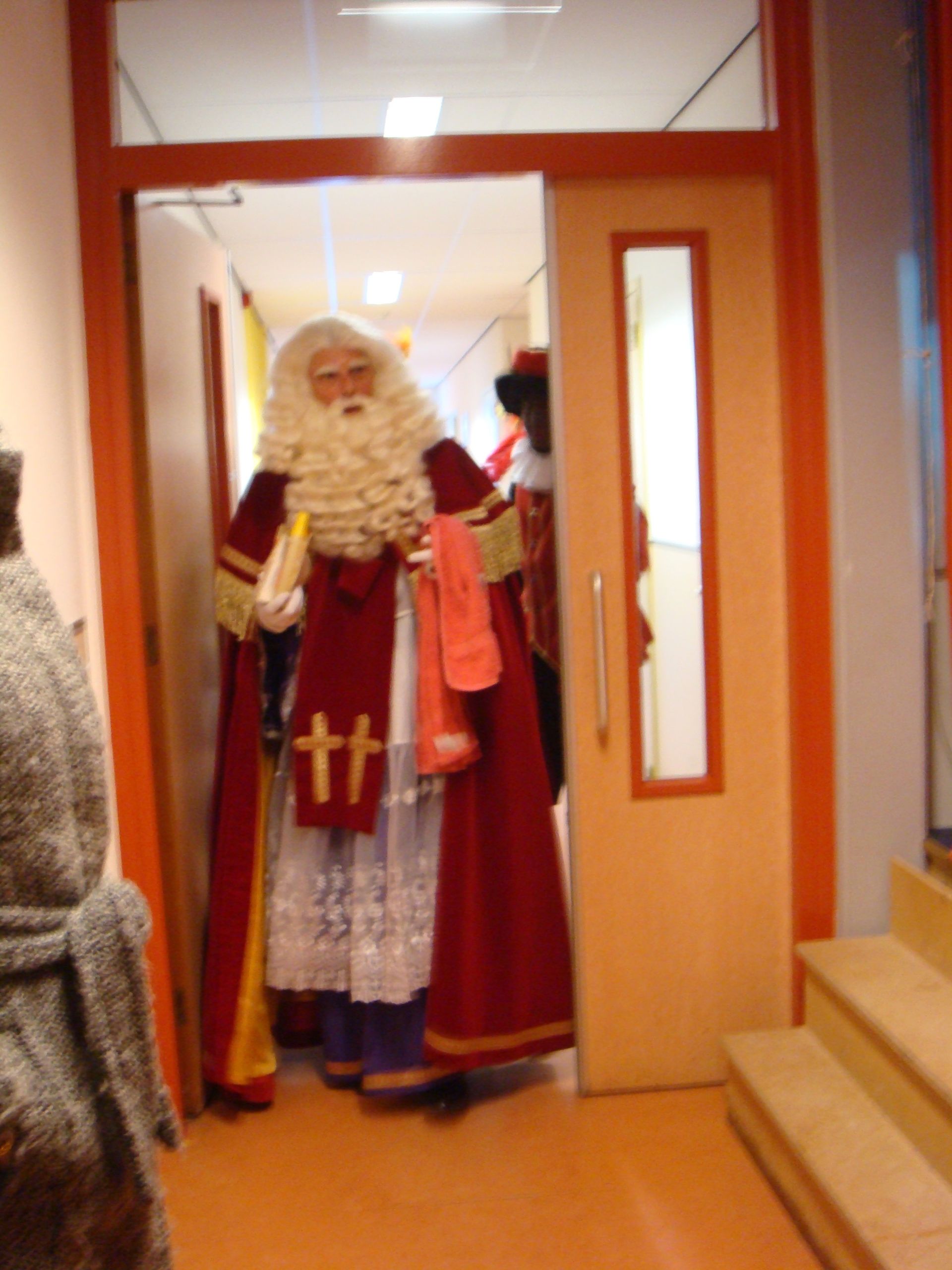 Sinterklaas, gekleed in traditionele rode gewaden en witte kanten kleding, loopt door een open deuropening, vergezeld door een assistent.