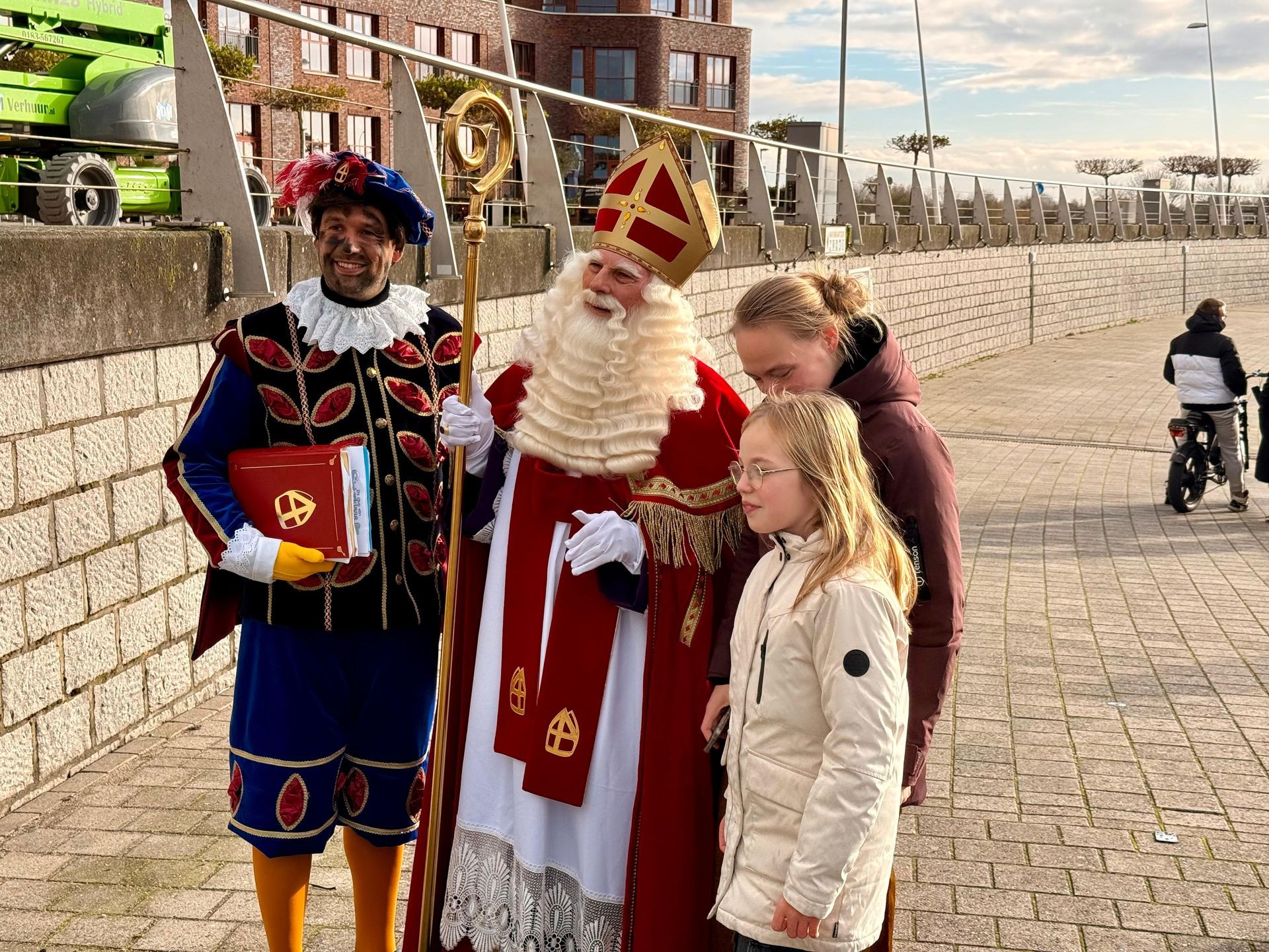 Sinterklaas en een helper in traditionele klederdracht staan ​​buiten op een stenen pad en begroeten twee mensen.