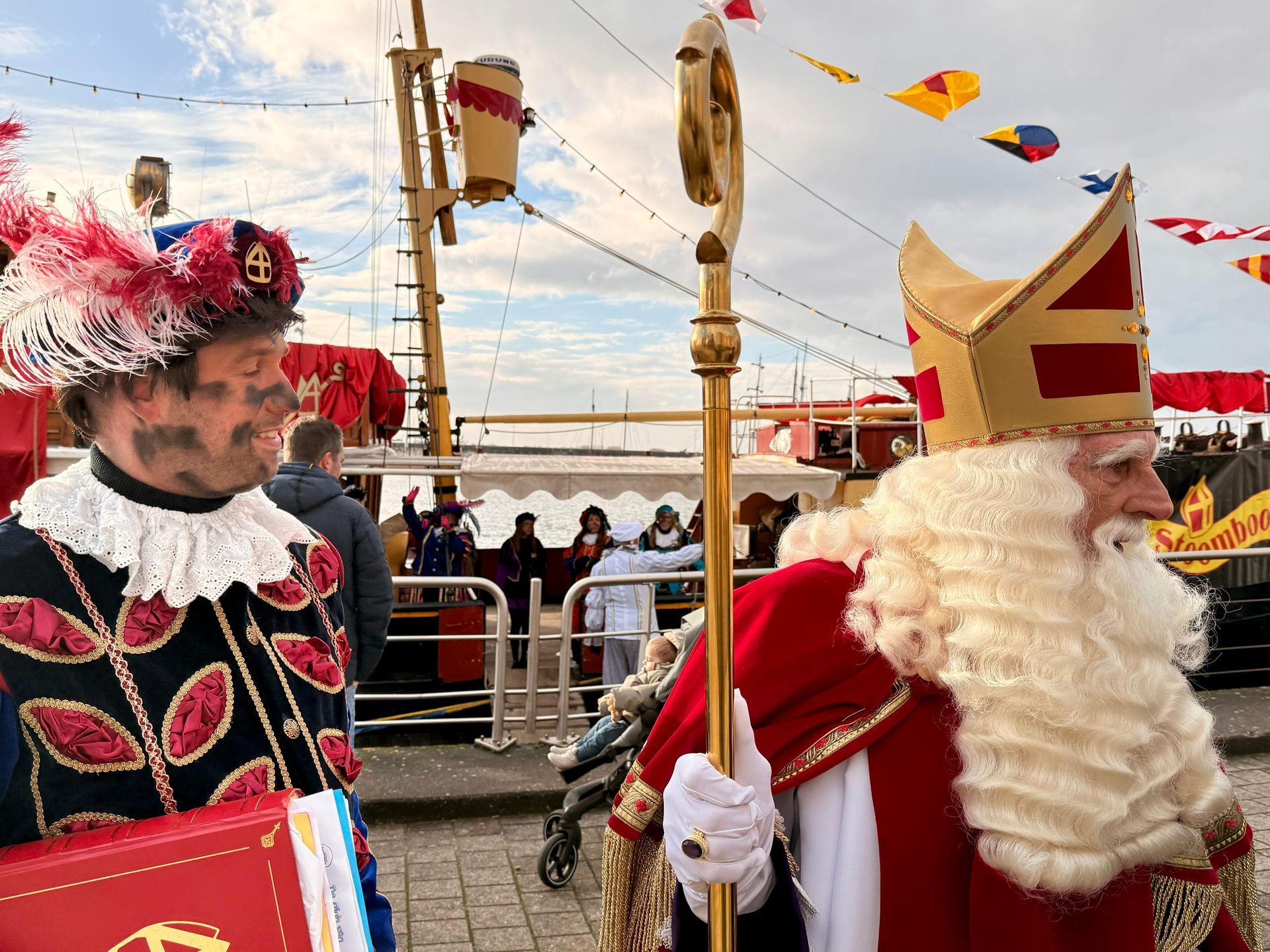 Sinterklaas met zijn mijter en staf, en Zwarte Piet, staan ​​samen buiten met een schip op de achtergrond.