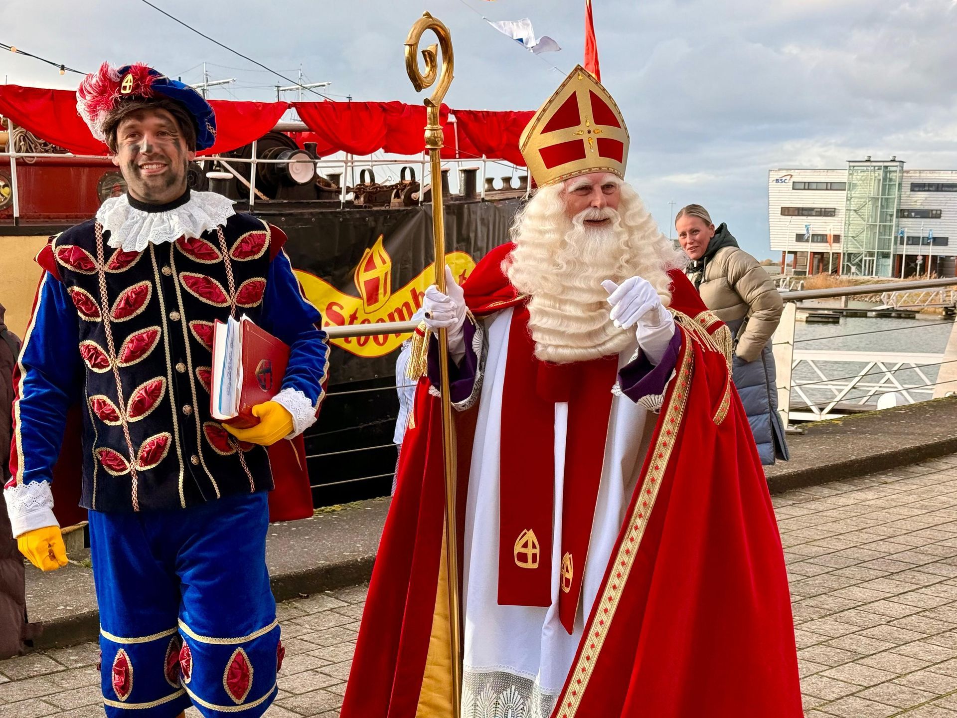 Sinterklaas en een metgezel in traditionele feestkleding staan ​​bij een haven, met een boot op de achtergrond.