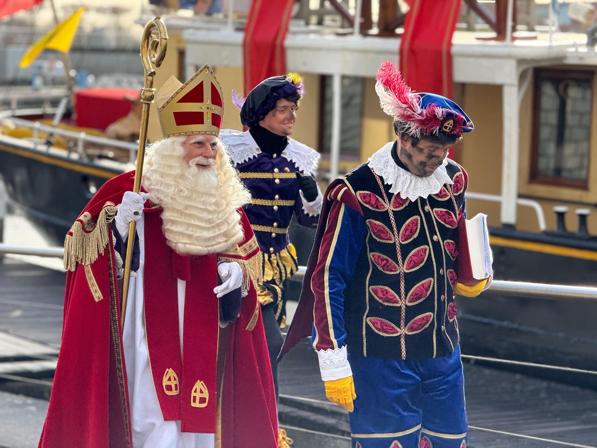 Sinterklaas met zijn gouden staf en twee helpers in uitbundige, kleurrijke kostuums in renaissancestijl loopt langs een boot.