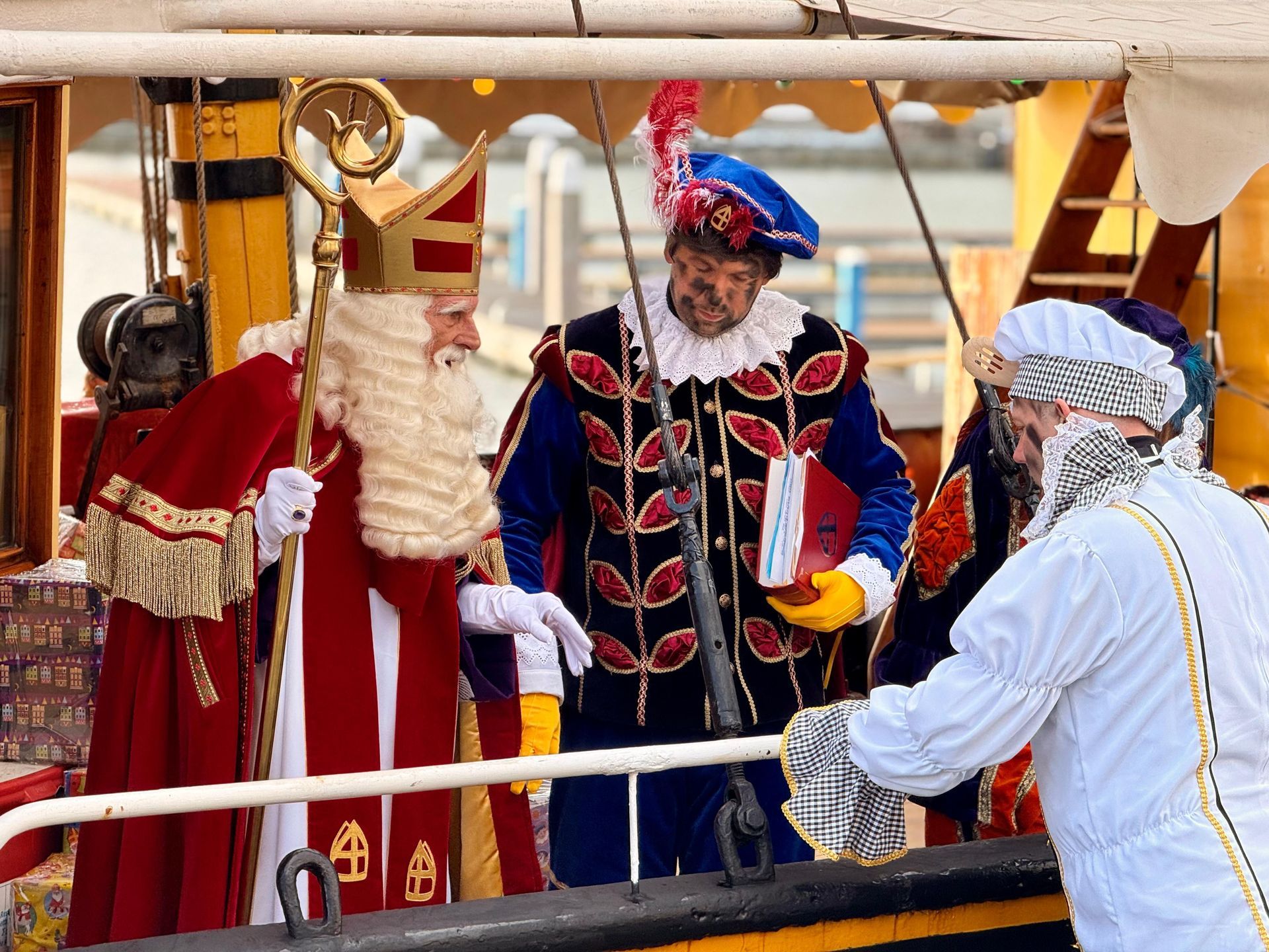 Sinterklaas staat met zijn staf en twee helpers op het dek van een houten schip tijdens de jaarlijkse Sinterklaas-aankomstviering.