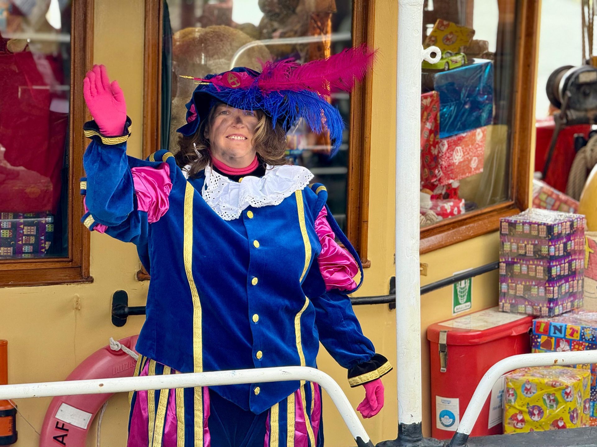 Een persoon in een blauw, roze en wit kostuum met een hoed met veren zwaait vanaf een bootdek dat versierd is met cadeaudozen.