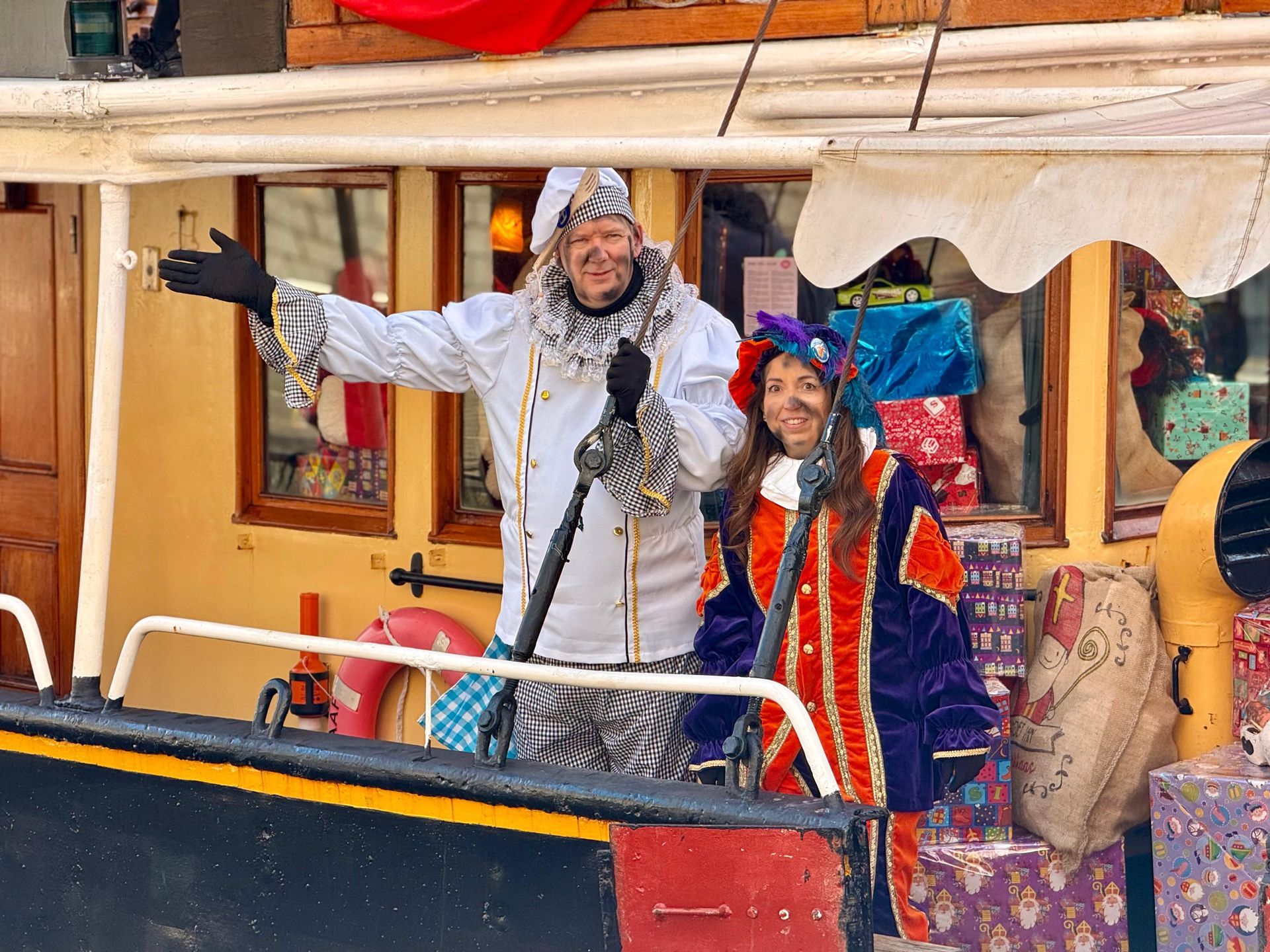 Twee mensen in kleurrijke carnavalskostuums zwaaien vanaf het dek van een boot die versierd is met ingepakte cadeaudozen.