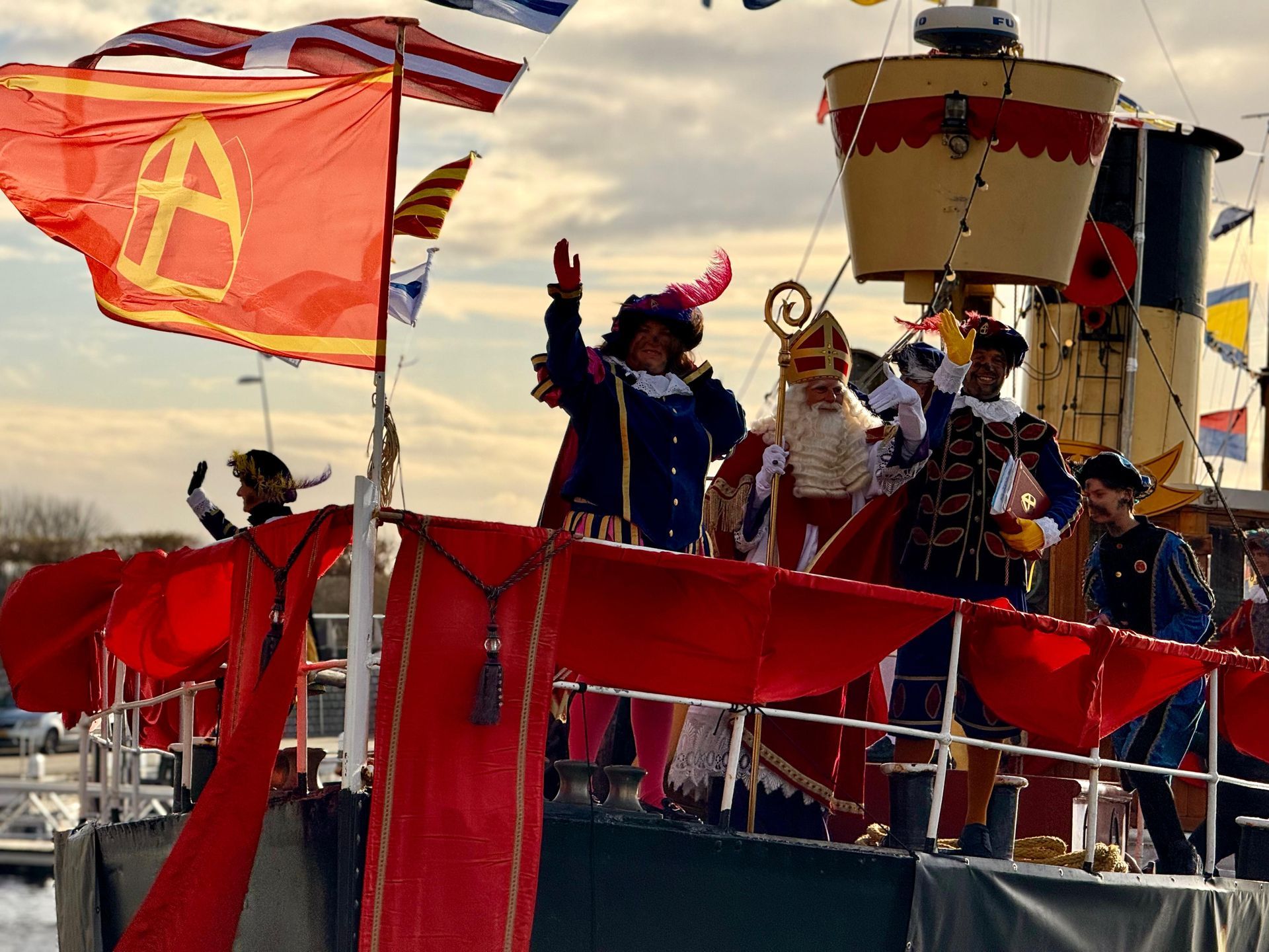 Een Sinterklaasparade op een boot met figuren in traditionele feestkleding die naar de menigte zwaaien.