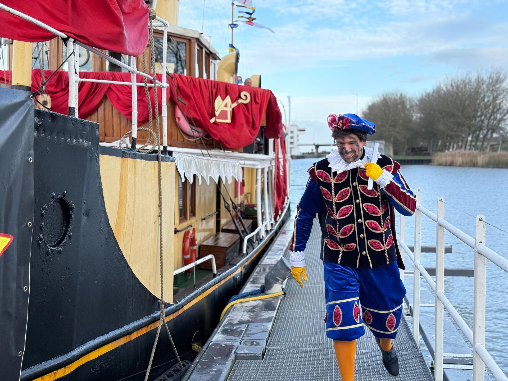 Een persoon in een kleurrijk, traditioneel Sinterklaaskostuum loopt over een metalen steiger naast een boot die versierd is met rode stof.