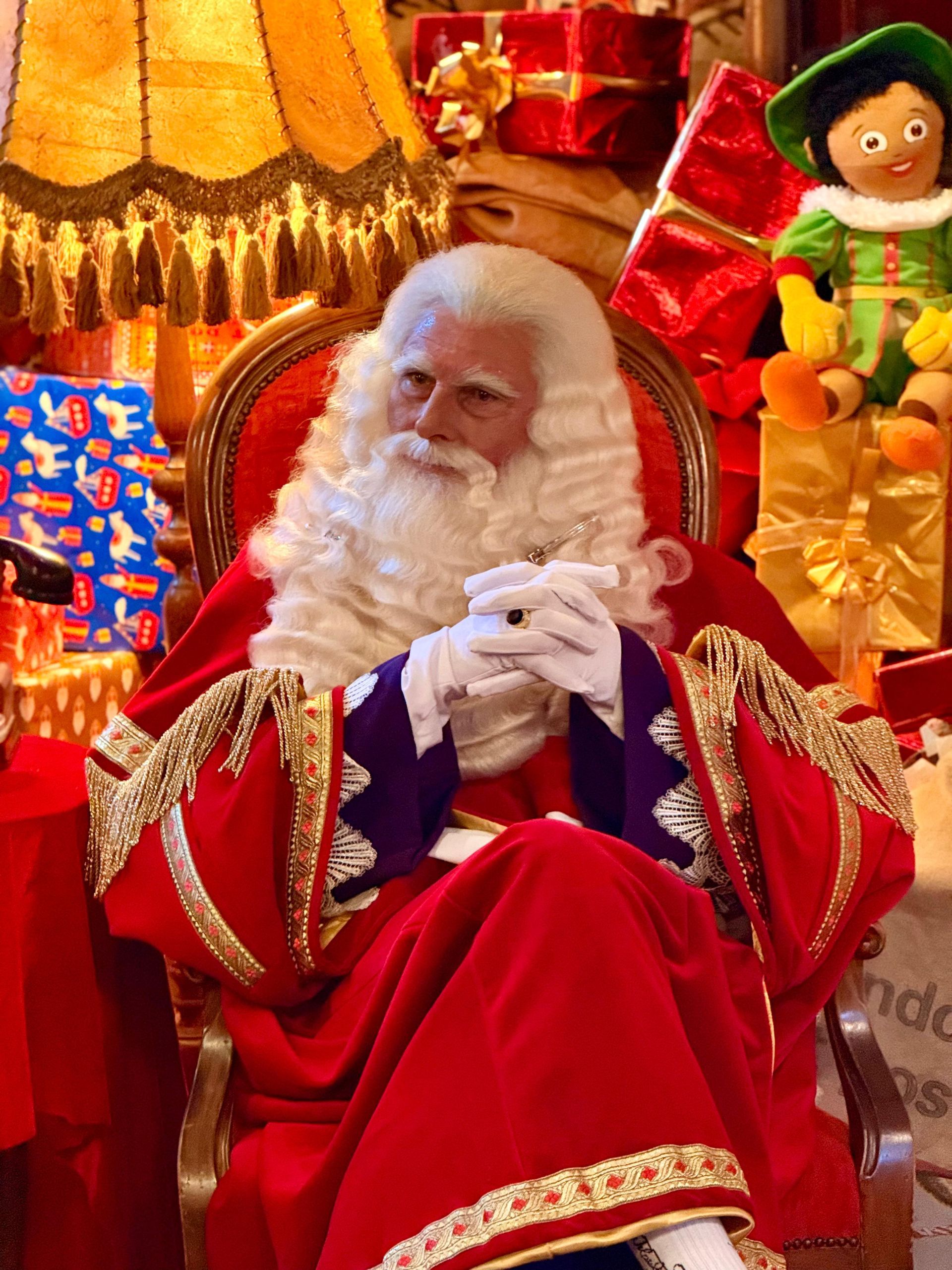 De Kerstman, gekleed in een rood fluwelen pak, zit op een stoel omringd door cadeaus en een feestelijke lamp.