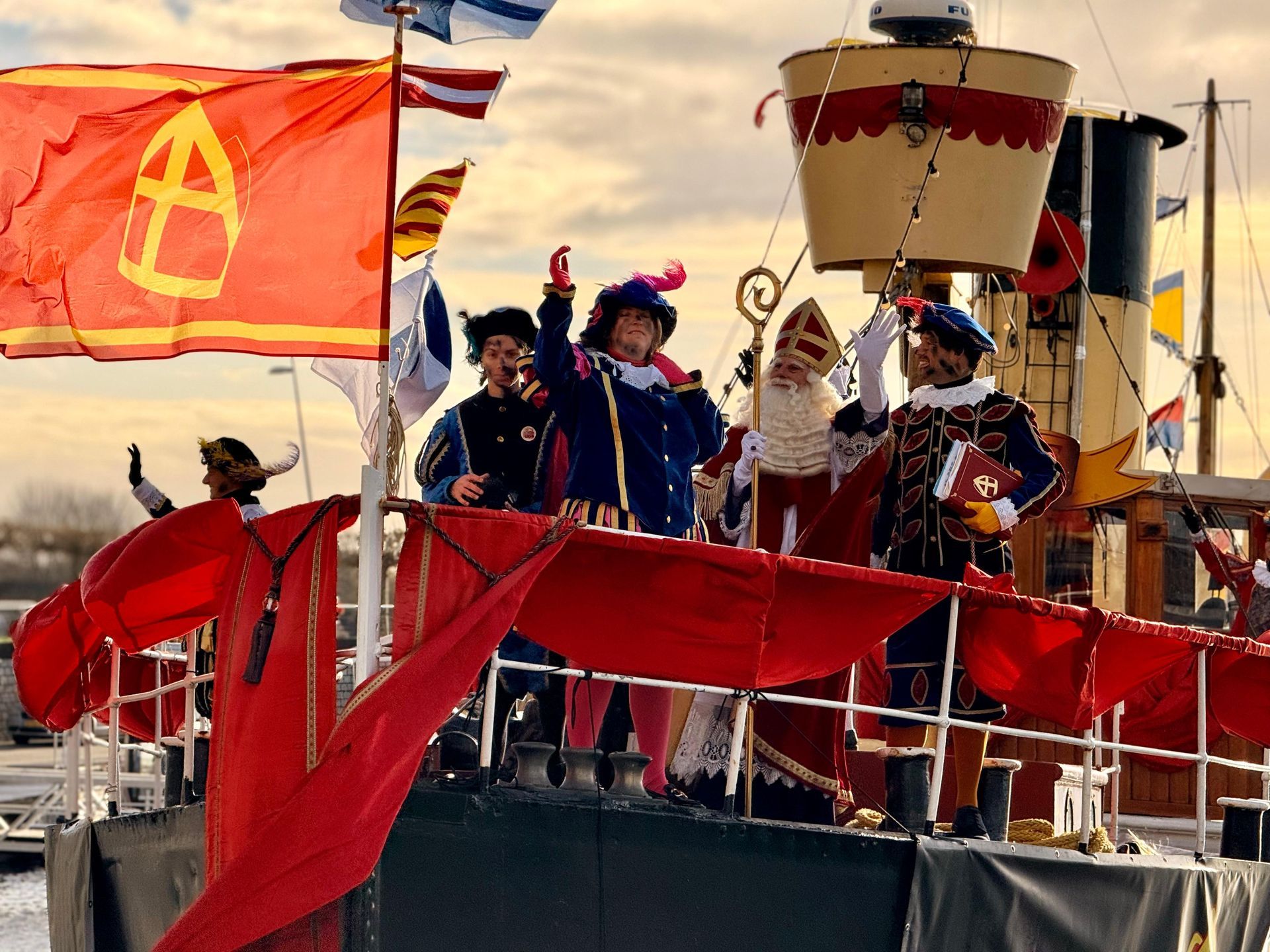 Sinterklaas en Pieten, gekleed in traditionele kostuums, zwaaien vanaf het dek van een met vlaggen versierde boot tijdens een feest.